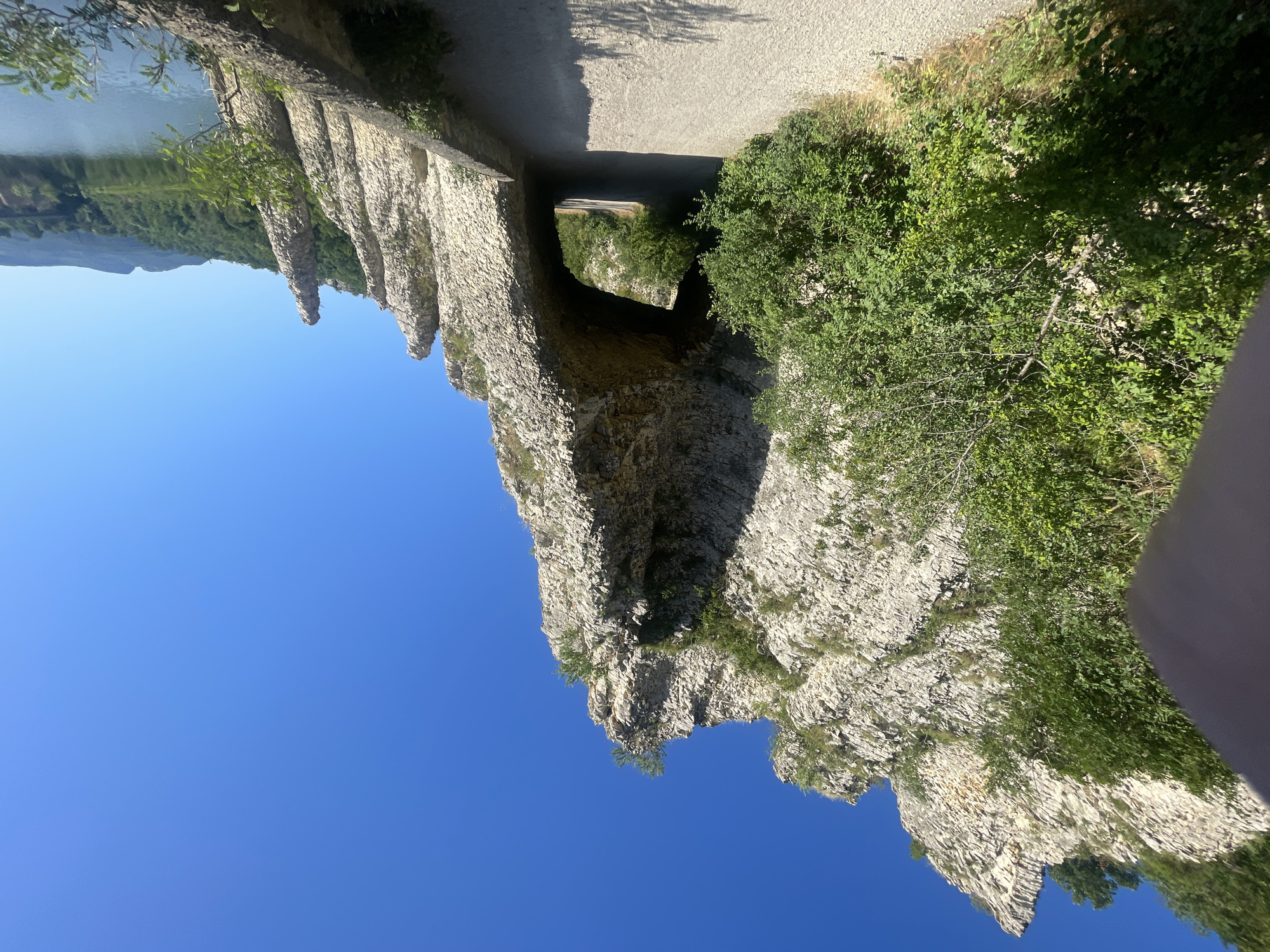 The image shows a road passing under a large natural rock arch with a clear blue sky above. The road is made of gravel and a stone wall runs along the right side. Lush green trees and vegetation are visible on the left, and the rock formations on the right appear jagged and pointed. In the background, a body of water is visible, reflecting the sky, with rolling green hills and mountains in the distance. This scene is a natural landmark located in Asparuhovo, Bulgaria, known as the "Stone Forest" or "Kamenni Gorat." There are no people visible in the image.