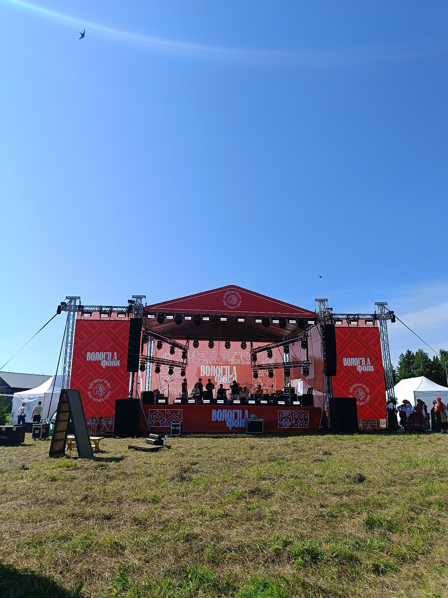In Semenkovo, Russia, a bright, sunny day unfolds with a large outdoor stage set up for a performance. The stage, draped in vibrant red banners featuring white Cyrillic text and intricate patterns, is the central focus. Two large speakers are mounted on either side of the stage's facade. A band of at least five musicians is visible on stage, performing. The ground in front of the stage is covered in green grass, with some areas appearing dry and patchy. To the left of the stage, white tents are set up, and a few people are gathered near them. To the right, more white tents and a cluster of people dressed in what appears to be traditional folk attire are visible, suggesting a cultural event or festival. A light pole with wires extends from the left side of the stage structure towards the sky. The sky is a clear, brilliant blue with a faint lens flare visible in the upper left quadrant. A bird is captured in flight against the blue expanse. The overall scene depicts an open-air cultural event in a rural or park-like setting.