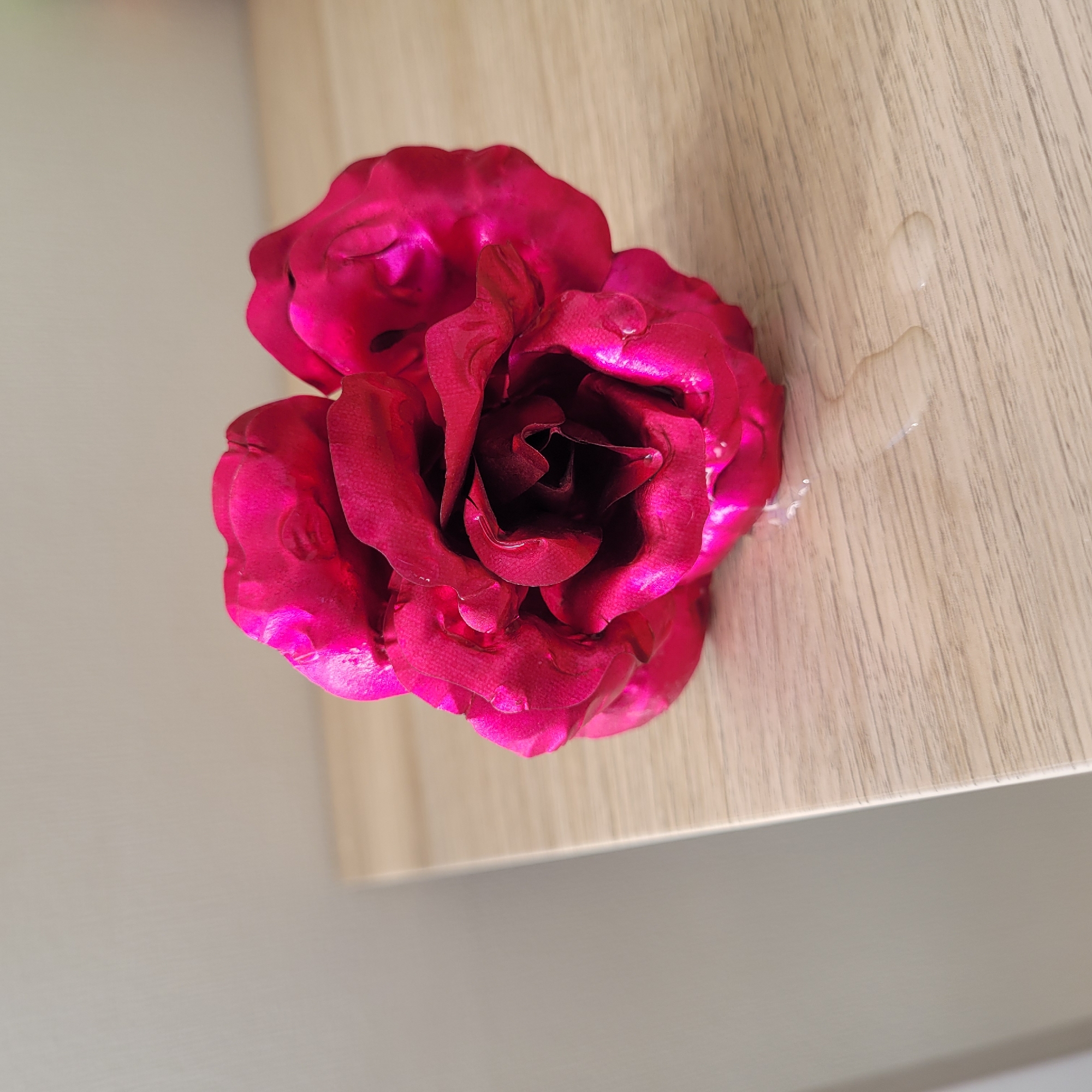 A close-up shot focuses on a bright pink rose, appearing somewhat artificial, resting on a light-colored wooden surface. The rose has multiple layers of petals, some of which are creased and slightly curled. Several small droplets of water are visible on the petals and scattered on the wooden surface in front of the rose. The background consists of a plain, light-colored wall. The overall lighting suggests an indoor setting, possibly on a table or shelf, with a shadow cast by the rose. There are no people or animals in the image. The environment is clean and uncluttered. The image does not provide any information about the location in the city of Jūrmala, Latvia.