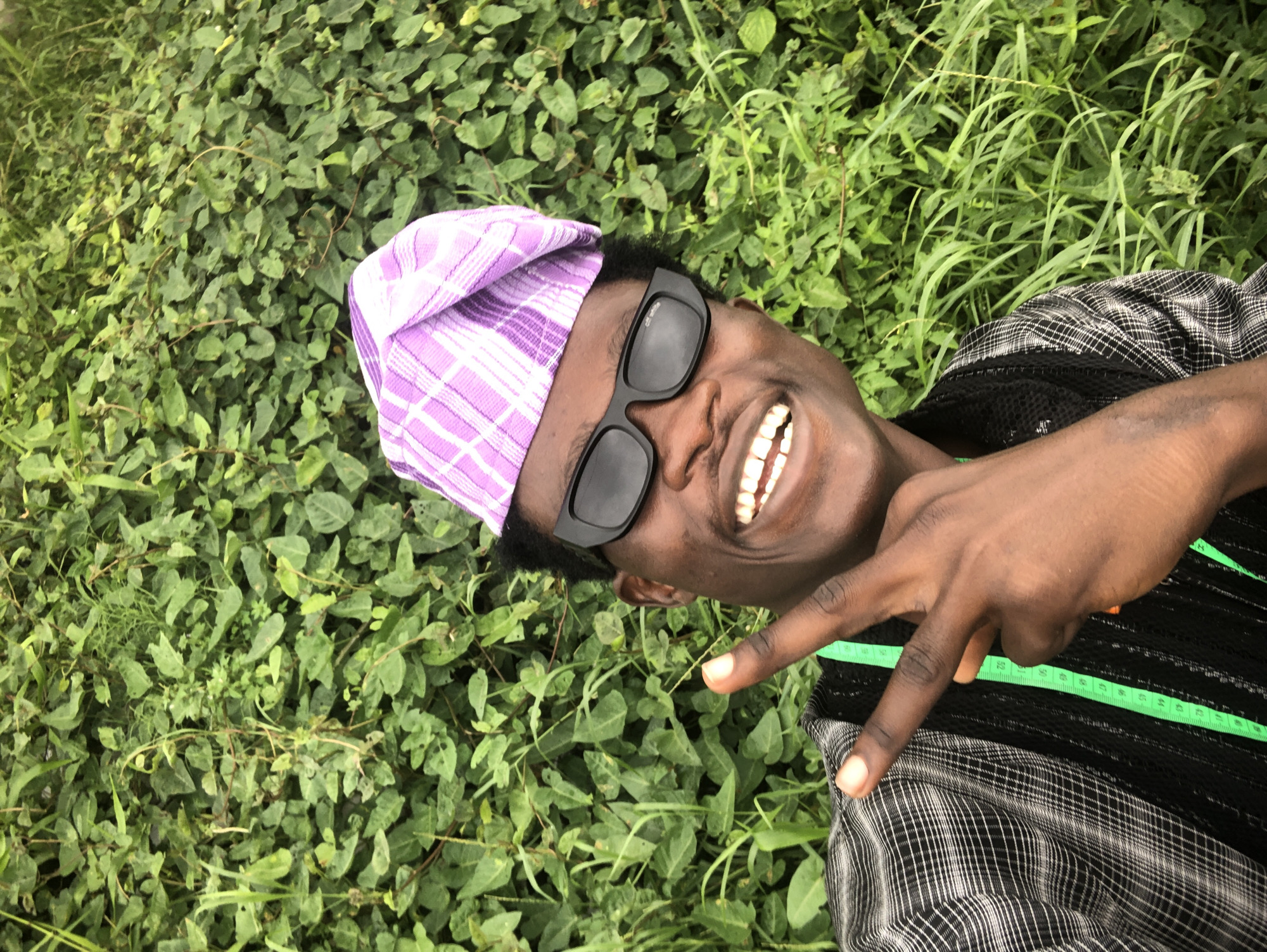 A man is pictured in a close-up, outdoor shot against a backdrop of lush green foliage. He is wearing a light purple and white checkered cap, black framed sunglasses, and a black and gray checkered shirt with black mesh detailing. His mouth is open in a wide smile, revealing his teeth. His right hand is raised, with two fingers extended in a peace sign. A green measuring tape is visible, draped over his shoulder and chest area. The overall impression is a candid and cheerful portrait. The specific location in Ado, Nigeria, is not discernible from the visual information provided in the image.