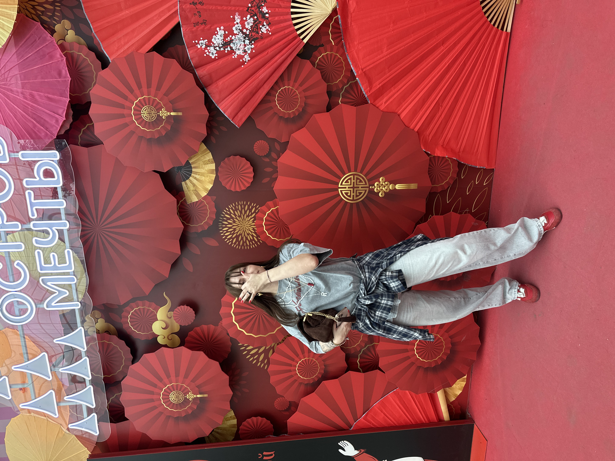 The image captures a young woman posing in an indoor setting against a vibrant red and gold decorative backdrop. The scene is dominated by numerous red paper fans and circular embellishments of varying sizes, many featuring intricate gold patterns, including stylized designs, geometric motifs, and traditional knots. Some fans also display white floral branches against the red. Smaller gold cloud shapes and abstract patterns are scattered across the dark red background.
The woman stands towards the center-right, wearing light-colored jeans, a grey t-shirt, and a blue and white plaid shirt tied around her waist. She has red sneakers and carries a small brown handbag. Her outfit is complemented by multiple pearl necklaces and rings. She poses with her hands raised, partially obscuring her face, possibly in a playful or modest gesture. Her fingernails are painted red.
On the far left, a large translucent sign with white and blue lettering in Russian is partially visible. The text reads "ОСТРО... МЕЧТЫ," which translates to "Island of Dreams" (ОСТРОВ МЕЧТЫ). The overall environment, with its abundant red and gold decorations, suggests a festive or celebratory atmosphere, possibly a public art installation or photo booth. Given the context of Moscow, Russia, this elaborate display likely serves as an eye-catching attraction.