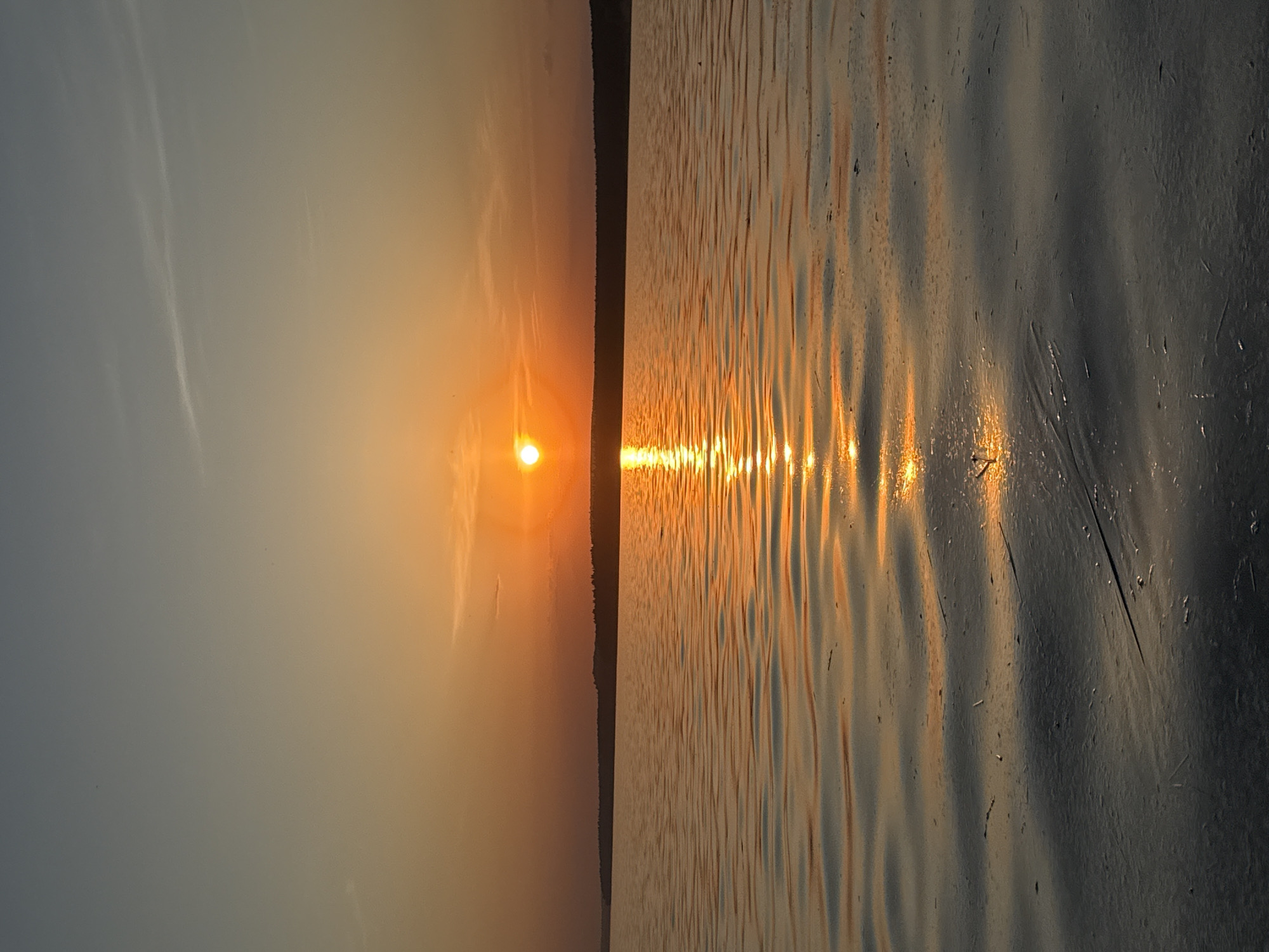 The image captures a sunset scene over a body of water, likely the Votkinsk Reservoir given the prompt's reference to Votkinsk, Russia. The sun is positioned near the horizon, casting a golden reflection on the water's surface. The reflection is striated due to ripples and waves on the water. A dark treeline is visible on the horizon. The sky transitions from a pale blue to orange-yellow near the sun. There are a few faint clouds visible in the sky. The water's surface appears partially frozen, with some ice formations.