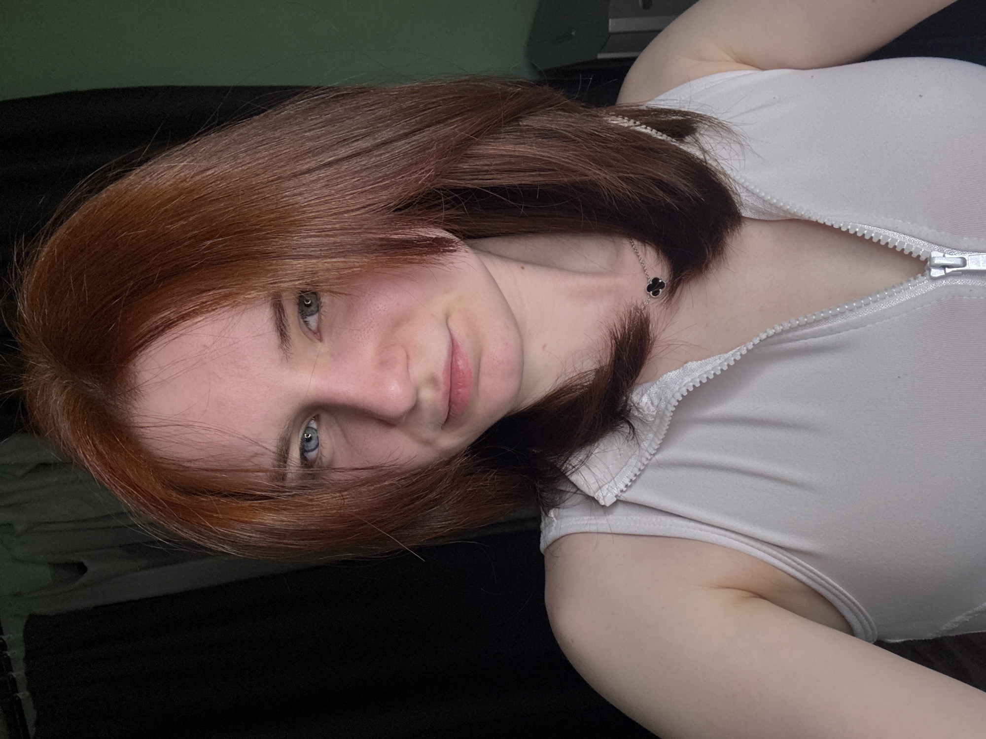 A woman with auburn hair, light skin, and blue eyes is angled towards the camera. She is wearing a light-colored top with a zipper partially undone at the collar. A necklace with a four-leaf clover pendant is visible. The background consists of a dark fabric or surface and an indistinct object, possibly electronic equipment, in the upper portion. No location in Vsevolozhsk, Russia is evident from the image.