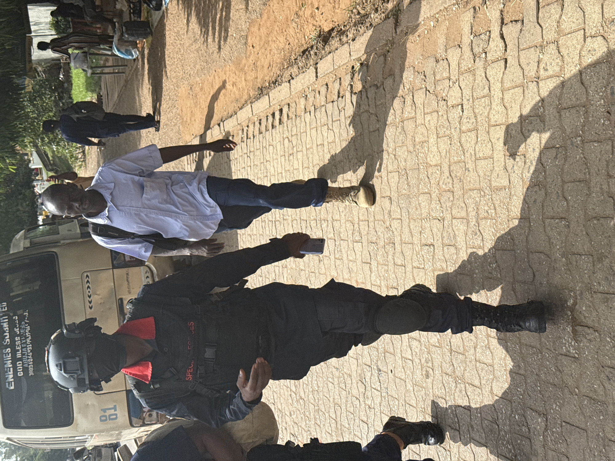 A daytime scene on a paved street in Abuja, Nigeria, depicts a man in civilian clothing being confronted by a Special Forces officer in full tactical gear. The officer is wearing a helmet, body armor, and boots and is holding a phone in his hand. A parked bus with the words "ENEMIES SUBMITTED - GOD BLESS JESUS - ABUJA, NIGERIA" and a phone number is in the background. There are other people in the background. The location is identifiable by the bus markings.