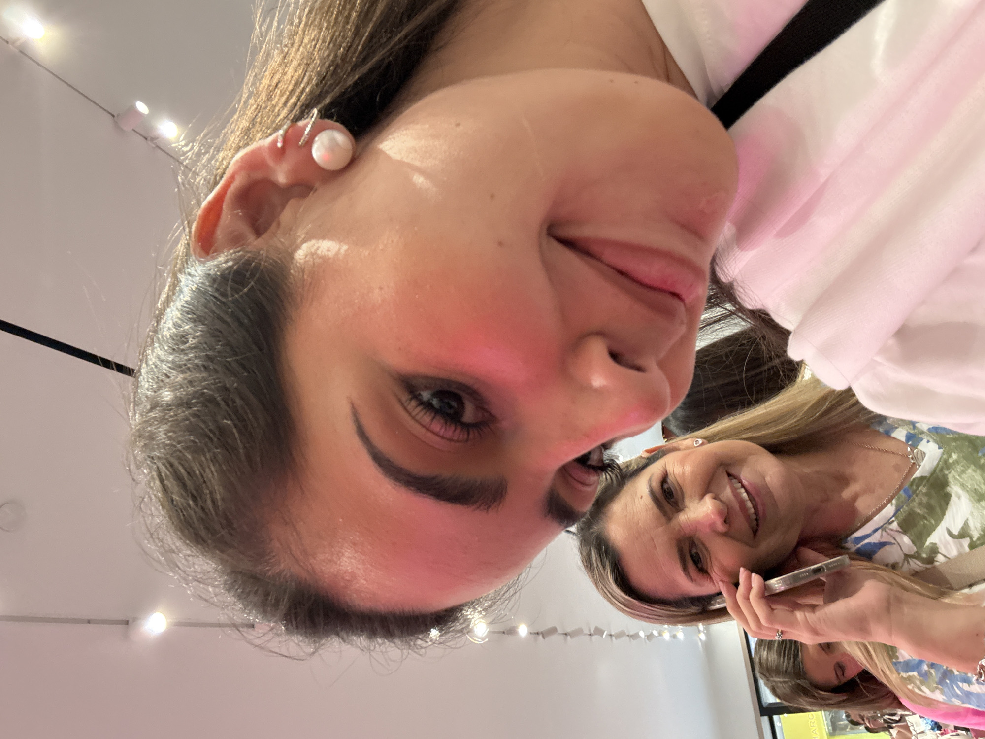 The image shows two women in an indoor setting. The foreground features a woman with dark hair and fair skin. She is wearing a white shirt and has a pearl earring. To her right, another woman with blonde hair is holding a smartphone. She is wearing a floral-patterned top and smiling. The background suggests a well-lit interior with a glimpse of what might be a shop window. The location appears to be a retail environment in San Antonio, United States of America.