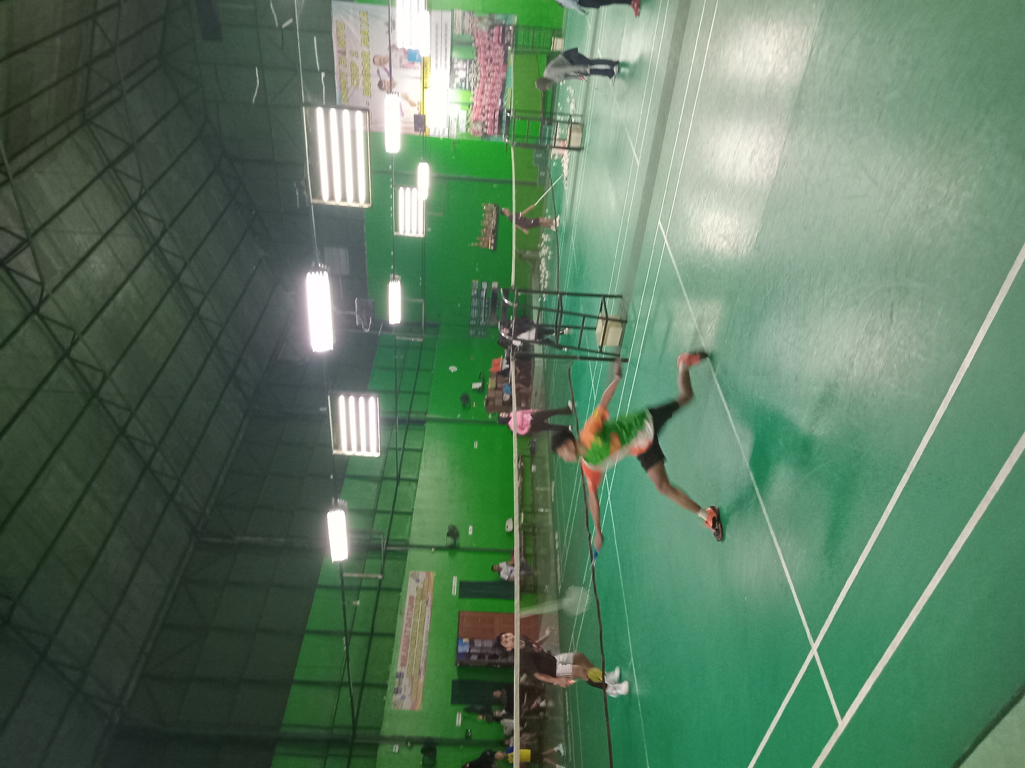 This image depicts an indoor badminton court with a green floor marked with white lines. Several individuals are present on the court, engaged in or observing a badminton match. In the foreground, a player in motion, wearing an orange and green shirt, is in the process of hitting a shuttlecock. Other players and spectators are visible in the background. The court is illuminated by overhead lighting, and banners are displayed on the walls, suggesting a tournament or event. The environment appears to be a sports facility. The image's metadata indicates it was taken in Bojong Gede, Indonesia.