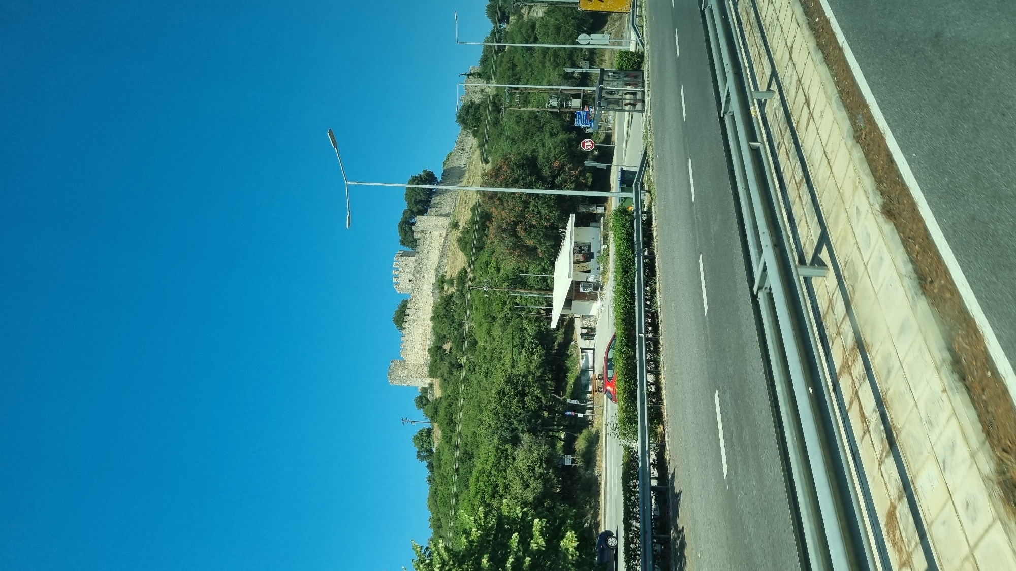 The image displays a clear, bright blue sky above a verdant hillside topped with a stone fortress. A paved road runs horizontally across the foreground, with a metal guardrail separating it from a sidewalk and a strip of vegetation. Several vehicles are visible on the road, including a red car and a dark blue car. Adjacent to the road, a small, white, open-air structure with a roof is situated on the hillside, possibly a bus stop or a small kiosk. Streetlights are positioned along the road, and the fortress on the hilltop features multiple turrets and crenellated walls, suggesting historical significance. The environment appears to be a sunny day, with ample greenery surrounding the fortress and the roadside. The scene is located in Neos Panteleimonas, Greece, near Platamonas Castle.