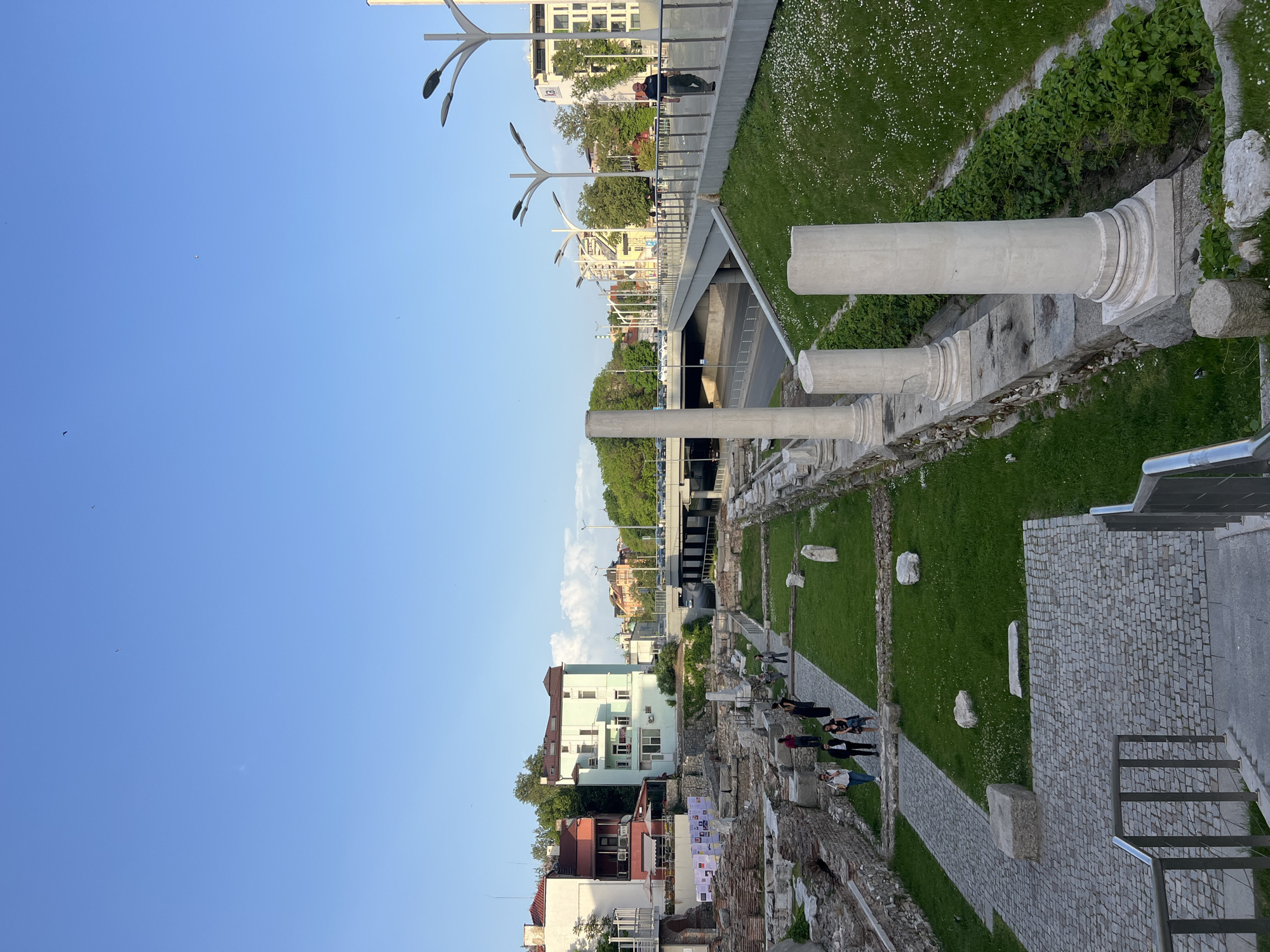 The image shows the ancient Roman Stadium in Plovdiv, Bulgaria. The ruins include parts of the stadium's seating and walls, and several marble columns. A paved walkway runs alongside the ruins, with grass and various other greenery. Several people are standing and walking along the walkway, seemingly observing the ruins. In the background, modern buildings and a road overpass are visible, showing the integration of the historical site into the modern city. The sky is clear and blue. Street lamps are visible above the highway.