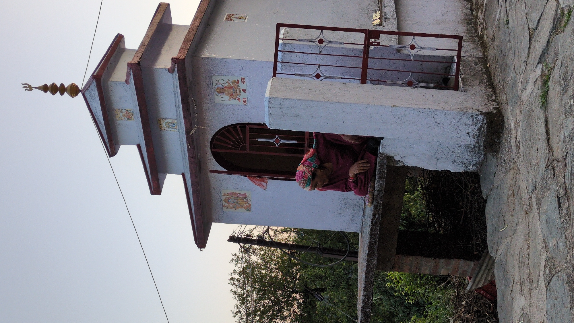 The image depicts a small Hindu temple or shrine in Haini, India, with an elderly woman observing her surroundings. The woman, dressed in a maroon garment and a multi-colored head covering, leans out from an arched opening or window in the temple, resting her arm on a ledge. She wears rings on her fingers and bracelets on her wrist.
The temple is a simple, white-painted structure with red trim and a tiered roof culminating in a golden trishul (trident) finial. Several religious images, likely posters or stickers, adorn the temple walls. Above one such image, the Devanagari script "जय श्री राम" (Jai Shri Ram) is clearly visible. A decorative metal railing, also red, is situated near another opening or balcony on the temple's upper level.
In the background, lush green trees and foliage are visible, suggesting a natural or semi-rural setting. To the right of the temple, a robust stone wall runs vertically through the frame. The sky is clear and bright, indicating either early morning or late afternoon with soft, diffused lighting. The overall scene portrays a tranquil, everyday moment at a local religious site.