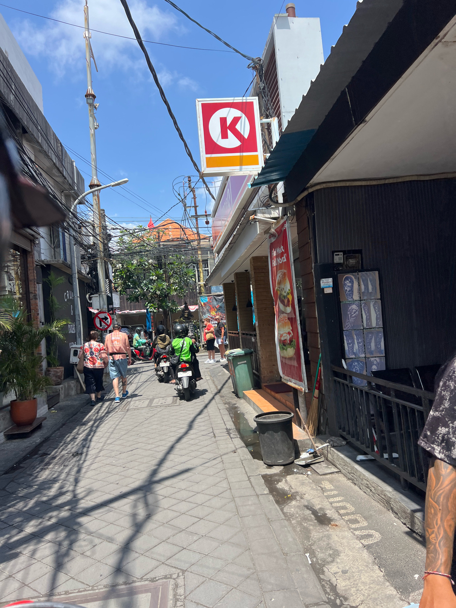 A low-angle shot captures a narrow, sun-drenched street in Kuta, Indonesia, on a bright, clear day. The street is paved with grey interlocking bricks, marked with subtle white lines. Buildings line both sides of the street. On the left, a multi-story building with dark facades and potted plants is visible, with a sign reading "CRU & GROSS." Further down, a red and white circular sign with a red "K" logo for Circle K hangs from a pole. On the right, a dark-colored building with a black corrugated metal facade has a metal railing in front. Attached to the building's facade is a large vertical red banner displaying images of two hamburgers. Next to it, a shelf holds several framed images of tattoos.
Several people are present. Two individuals, one in a patterned coral shirt and blue shorts, the other in a black top and pants, walk away from the camera on the left sidewalk. Further down the street, three people are riding motorcycles. The rider in the middle wears a bright green jacket, and the person behind them wears a yellow jacket. Another rider on a motorcycle, wearing a black helmet, is partially visible ahead. Another person wearing a red t-shirt and white shorts stands near a building entrance on the right. A person's tattooed arm and shoulder are visible in the lower right corner of the frame.
The environment is characterized by dense overhead electrical wires crisscrossing the sky and a tall utility pole with attached lights. Shadows from the wires and buildings cast long, distinct lines across the street. Trash cans, a broom, and a shovel are also present on the right side of the street. The overall scene depicts a bustling urban environment with a mix of commercial establishments and pedestrian traffic.