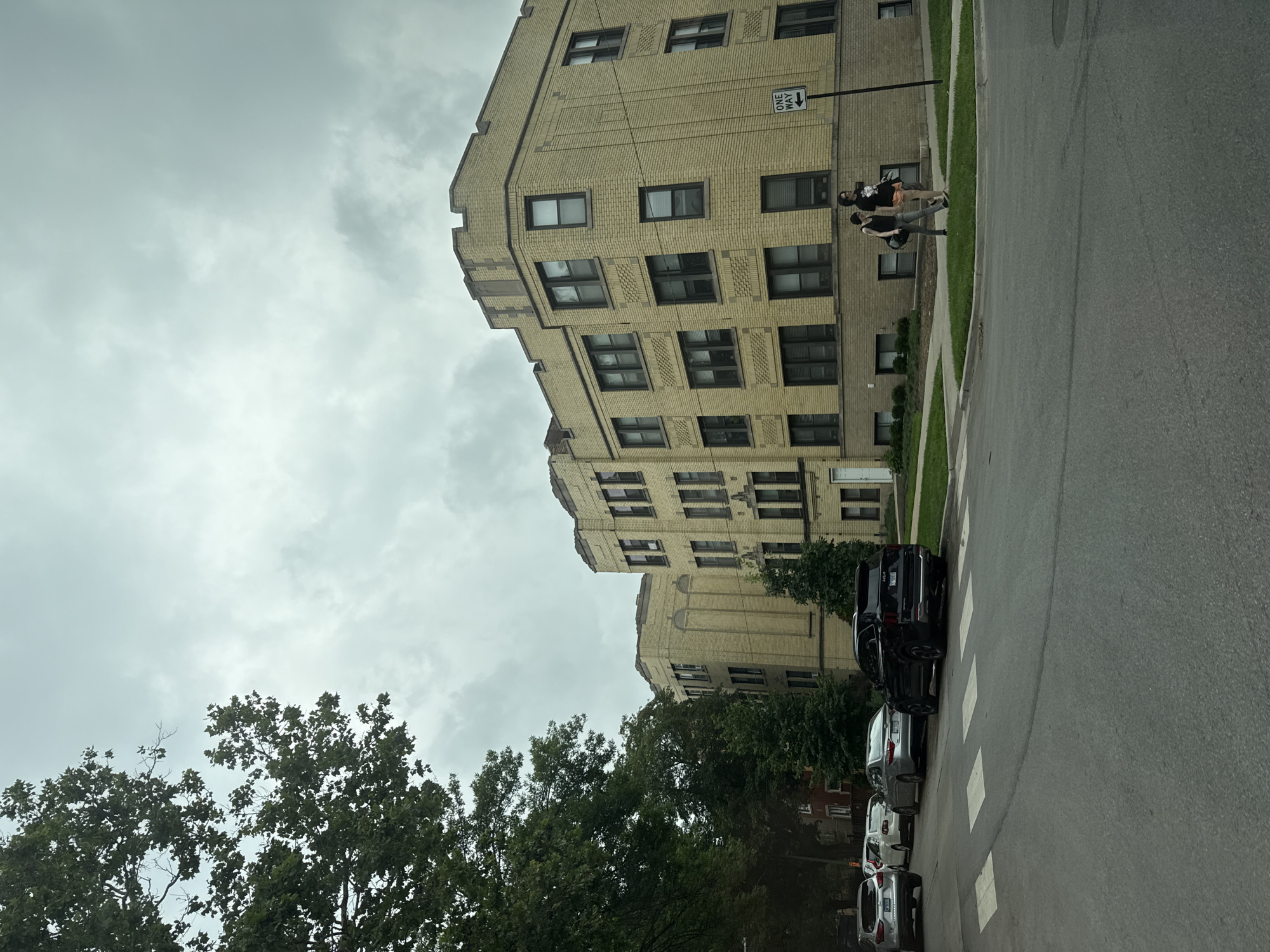 This image captures a street scene in Chicago, Illinois, USA, under an overcast sky. A yellow brick apartment building with dark-trimmed windows dominates the right side of the frame. Two young women are walking on the sidewalk in front of the building. A "One Way" street sign is visible next to them. Several cars are parked along the street, and a crosswalk is marked on the asphalt. Trees line the left side of the street, partially obscuring another building and the sky. The overall atmosphere suggests a typical urban neighborhood.