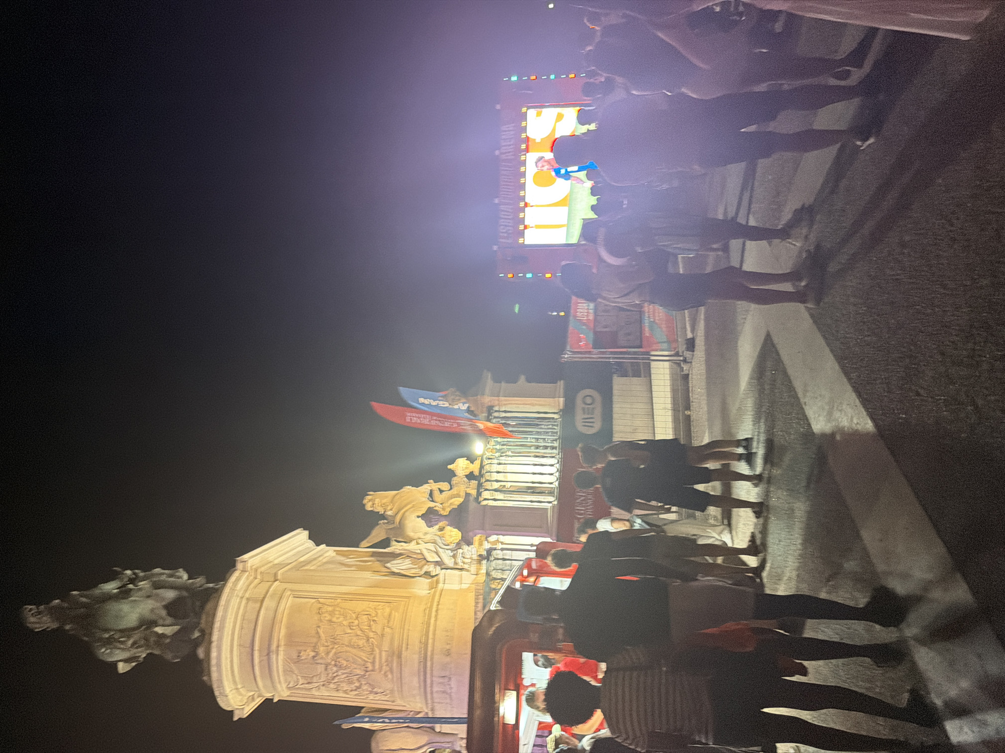 The image depicts a nighttime scene in Lisbon, Portugal, likely at Praça do Comércio, given the presence of the statue of King José I on horseback and the general ambiance of a public gathering. A large crowd of people is visible, standing in front of a large LED screen displaying an image of a football player and the text "LISBOA FOOTBALL ARENA". The screen is illuminated, casting a bright light on the surrounding area.
In the foreground, several individuals are standing, some facing the screen, others in profile. Their attire suggests a casual, outdoor event. To the left of the image, a red structure, possibly a food truck or kiosk, is partially visible. Behind the people, a large, ornate pedestal supports a bronze equestrian statue, illuminated by spotlights. The statue is detailed, with the horse in mid-stride and the rider holding a baton. The base of the pedestal features relief sculptures.
Red and blue flags are planted behind the statue, and a metal barrier with a banner containing the text "MEGANE THANDUR" is also present. The ground appears to be paved and wet, reflecting the lights from the screen and the surrounding structures. The overall atmosphere is one of a lively public event, likely a sports viewing or festival, taking place in a significant historical and public space in Lisbon. The sky is dark, indicating nighttime.