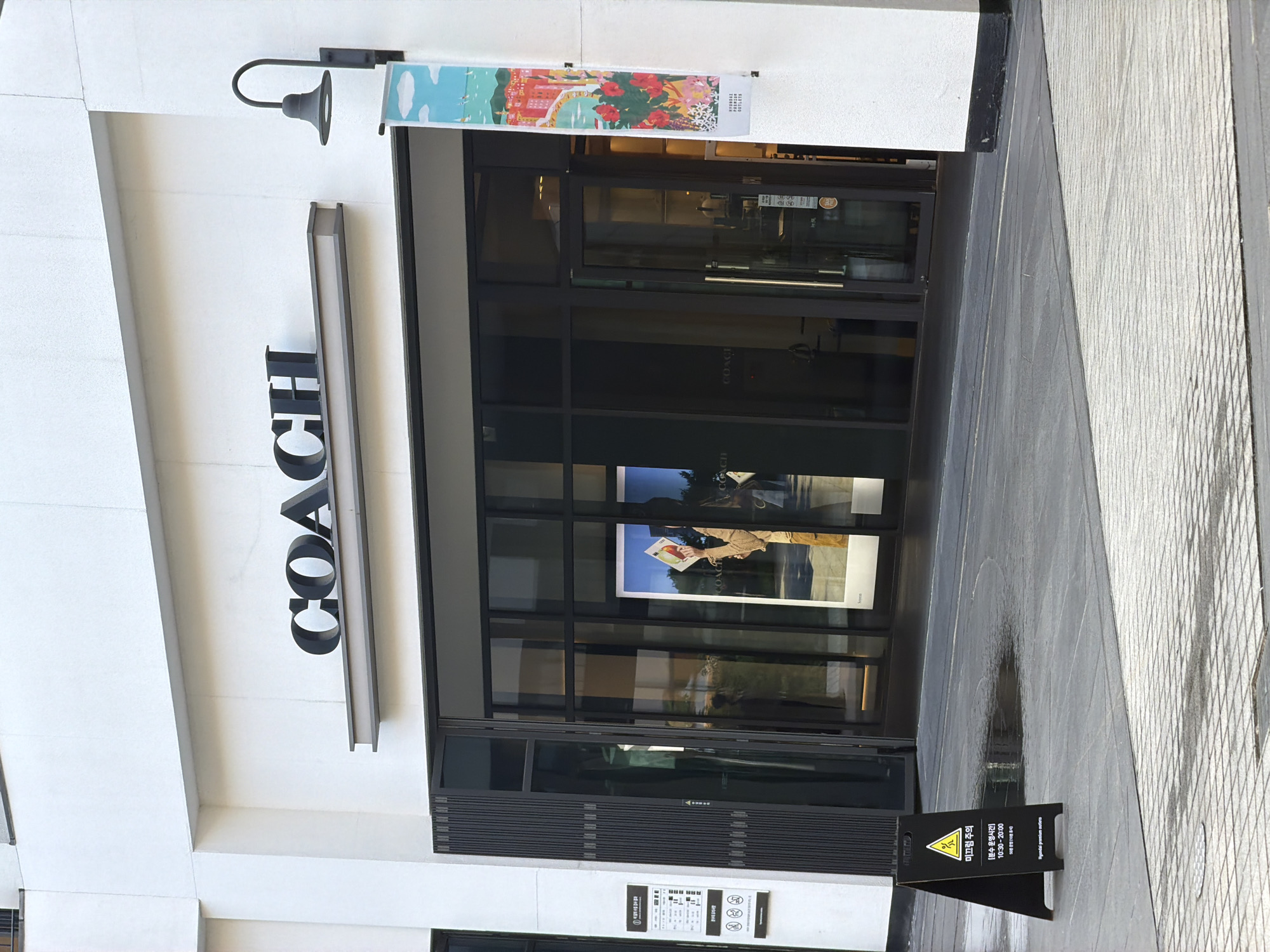 The image shows the entrance to a COACH store at a Hyundai Premium Outlets location in Incheon, South Korea. The store's large, dark-framed glass doors and windows are the central focus. The vertically oriented "COACH" lettering is prominently displayed on the white facade to the left of the entrance. Above the door, a colorful banner depicting a seaside town with flowers hangs. Inside the store, visible through the glass, are mannequins or displays showcasing clothing and accessories. A "Wet Floor" warning sign is placed on the patterned tiled ground to the right of the entrance, suggesting it may have recently rained or there's a water feature nearby. The lighting suggests it is daytime.