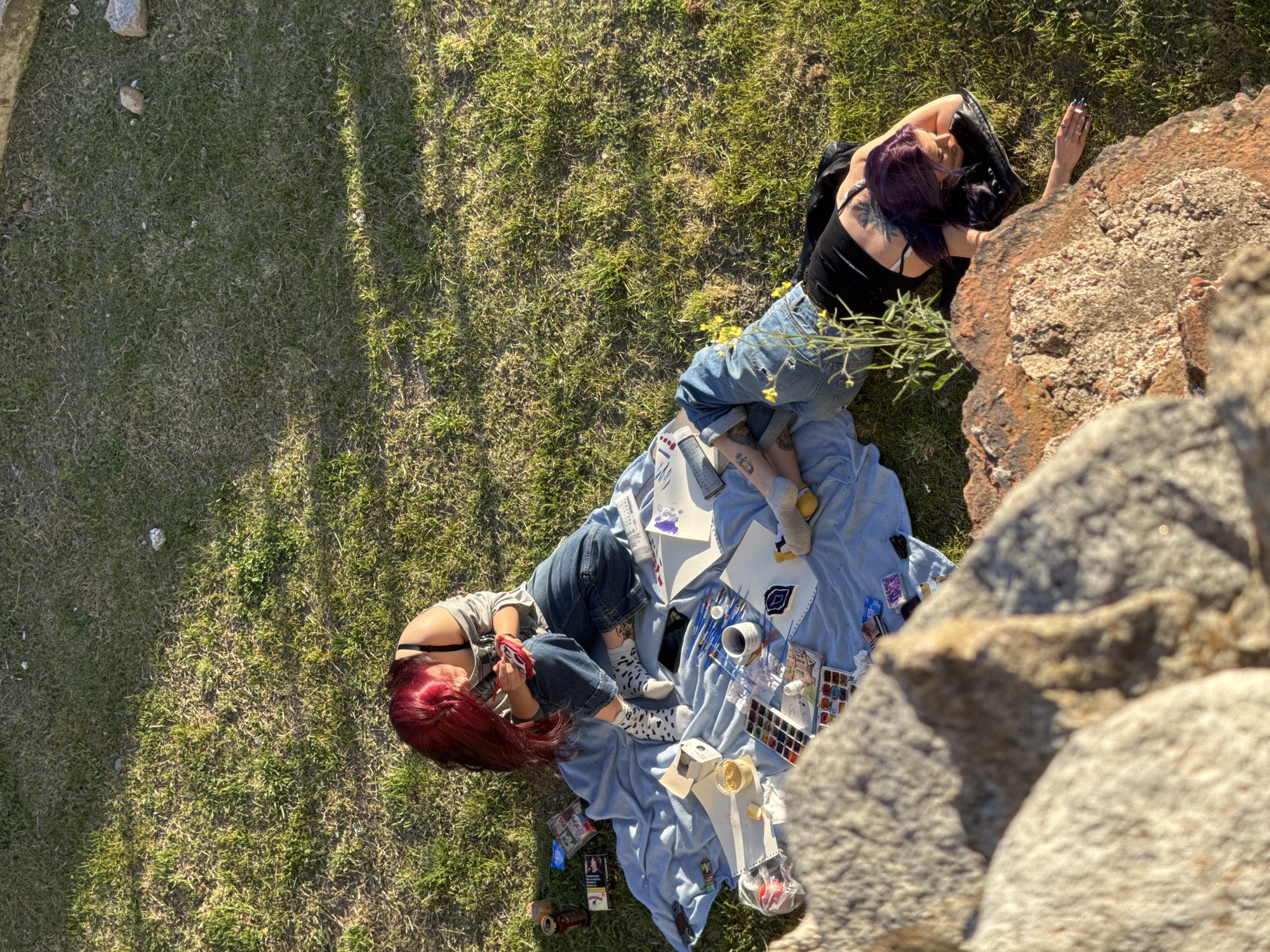 Two women lie on a blue blanket spread out on grass. One woman has long, red hair and wears a gray top and jeans. She looks at a red object in her hand. The other woman has dark hair with purple streaks, wears a black tank top and jeans, and her hand rests on a large rock. Paint supplies, including paintbrushes, paints, paper, and a cup, are arranged on the blanket. Grass surrounds the blanket on one side, and a large rock formation is on the other. The location is in Plovdiv, Bulgaria.