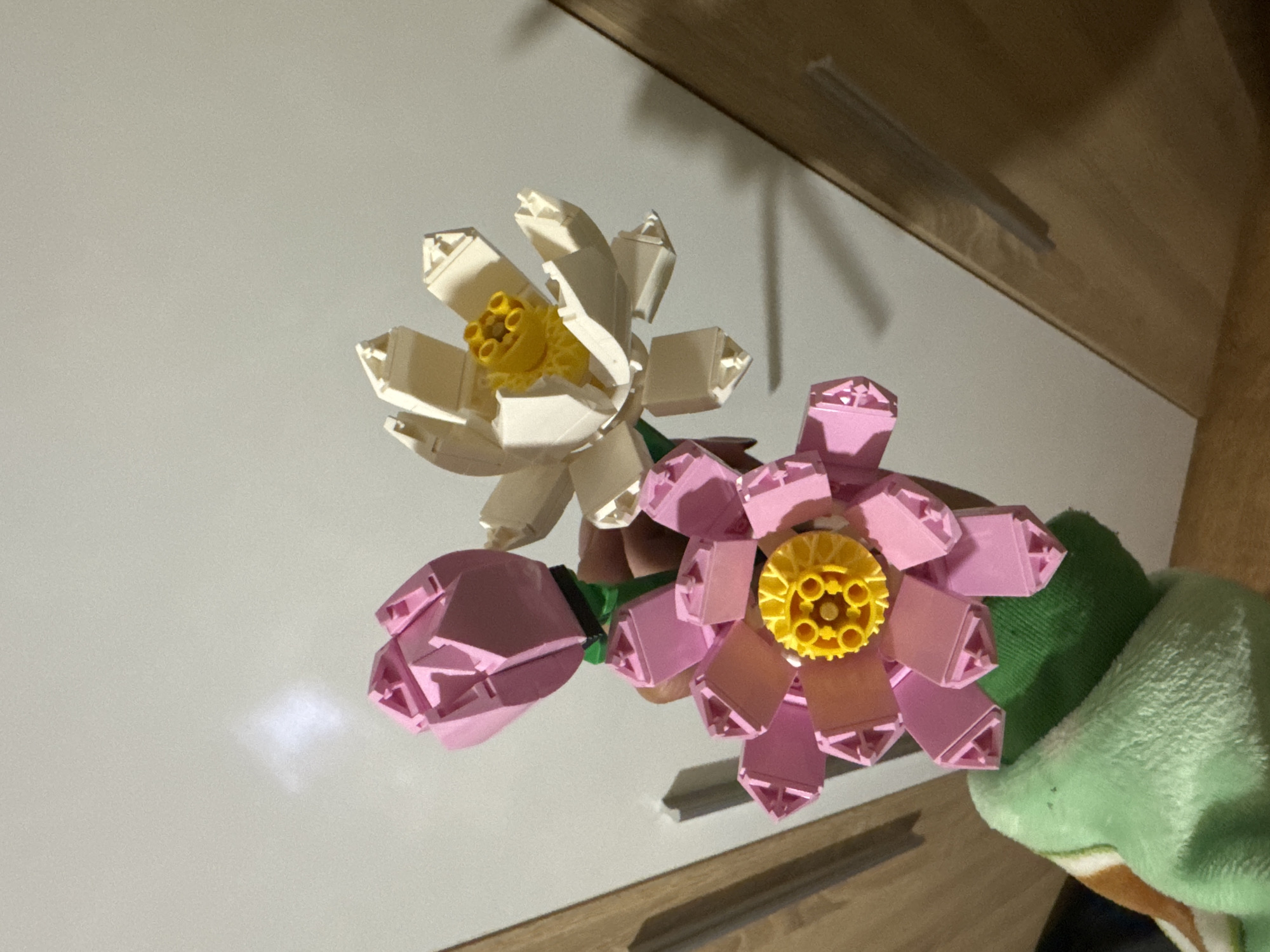 The image shows a bouquet of three Lego lotus flowers. One flower is white, one is pink, and the third is a pink bud. The flowers have yellow centers. They are resting on a white surface that appears to be a table or shelf. The background includes a wooden surface, possibly a cabinet or wall. There is also a light green cloth visible to the right of the flowers. The location is not discernible from this image.
