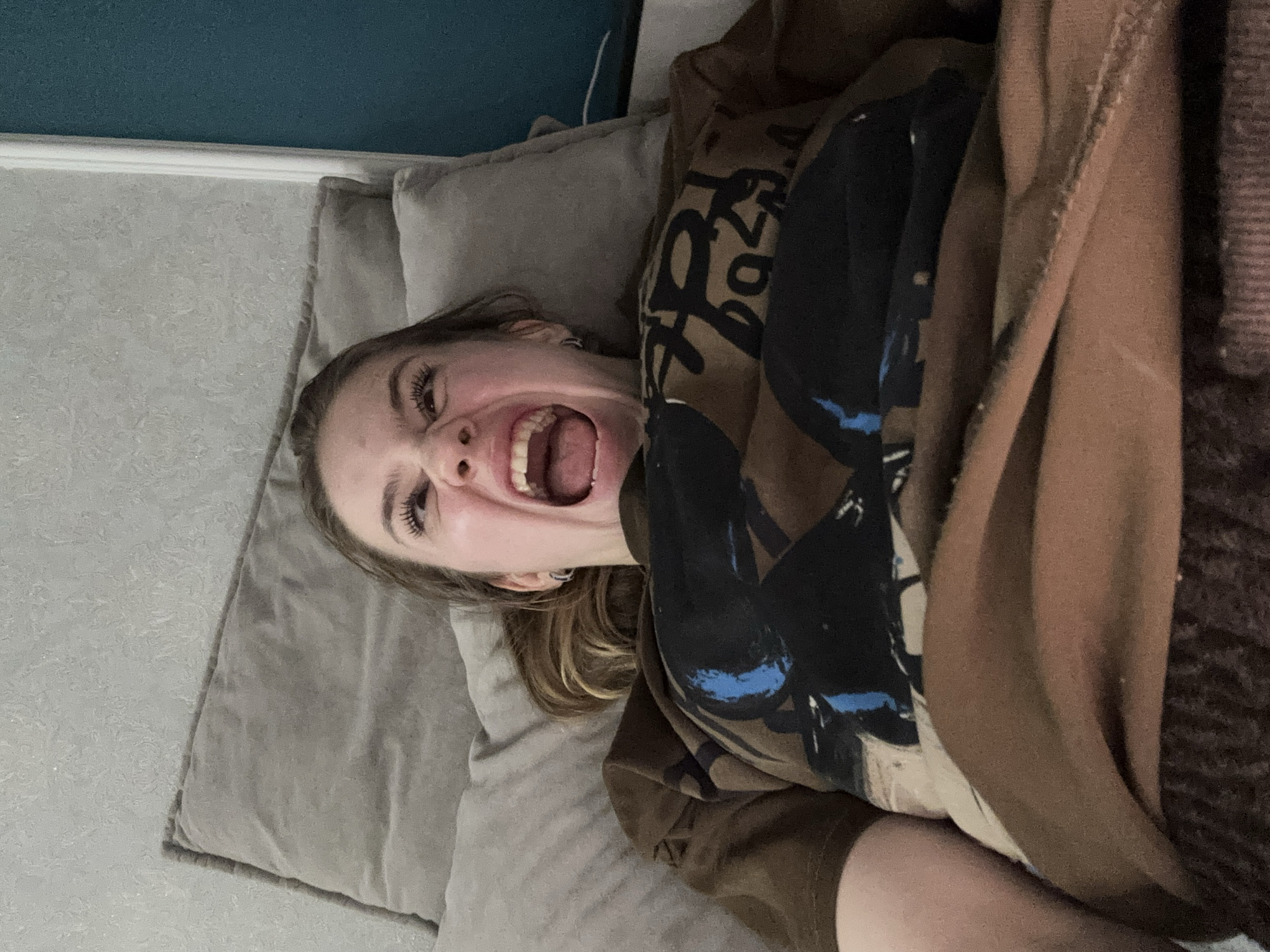 A young woman with blonde hair is lying down on her back on a bed. She is wearing a brown t-shirt with a black and blue graphic design. Her mouth is wide open in a scream or a laugh, and her eyes are squeezed shut. She appears to be in a bedroom, with pillows and a wall visible in the background. The lighting is dim, suggesting it may be nighttime or an overcast day. The image is taken from a high angle, looking down at the woman. The environment does not contain any specific visual cues that would indicate its location in Sinyavino, Russia.