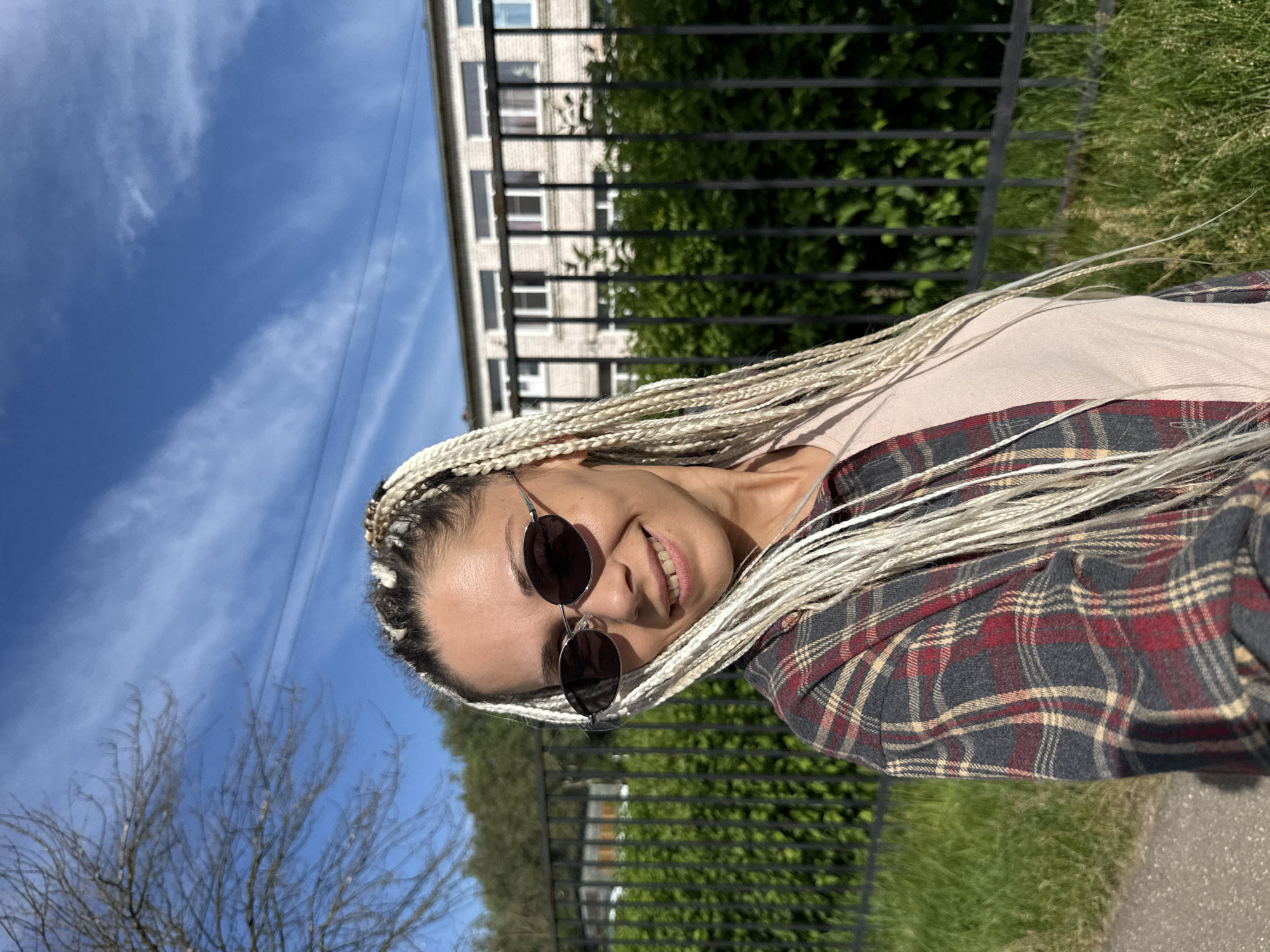 A woman with blonde braided hair is standing outdoors in front of a building and a fence. She is wearing sunglasses and a plaid shirt over a light-colored t-shirt. The background shows a building with windows, green bushes behind a black metal fence, and a blue sky with clouds. A bare tree branch is visible to the left. The location is identified as Rusanovo, Russia.