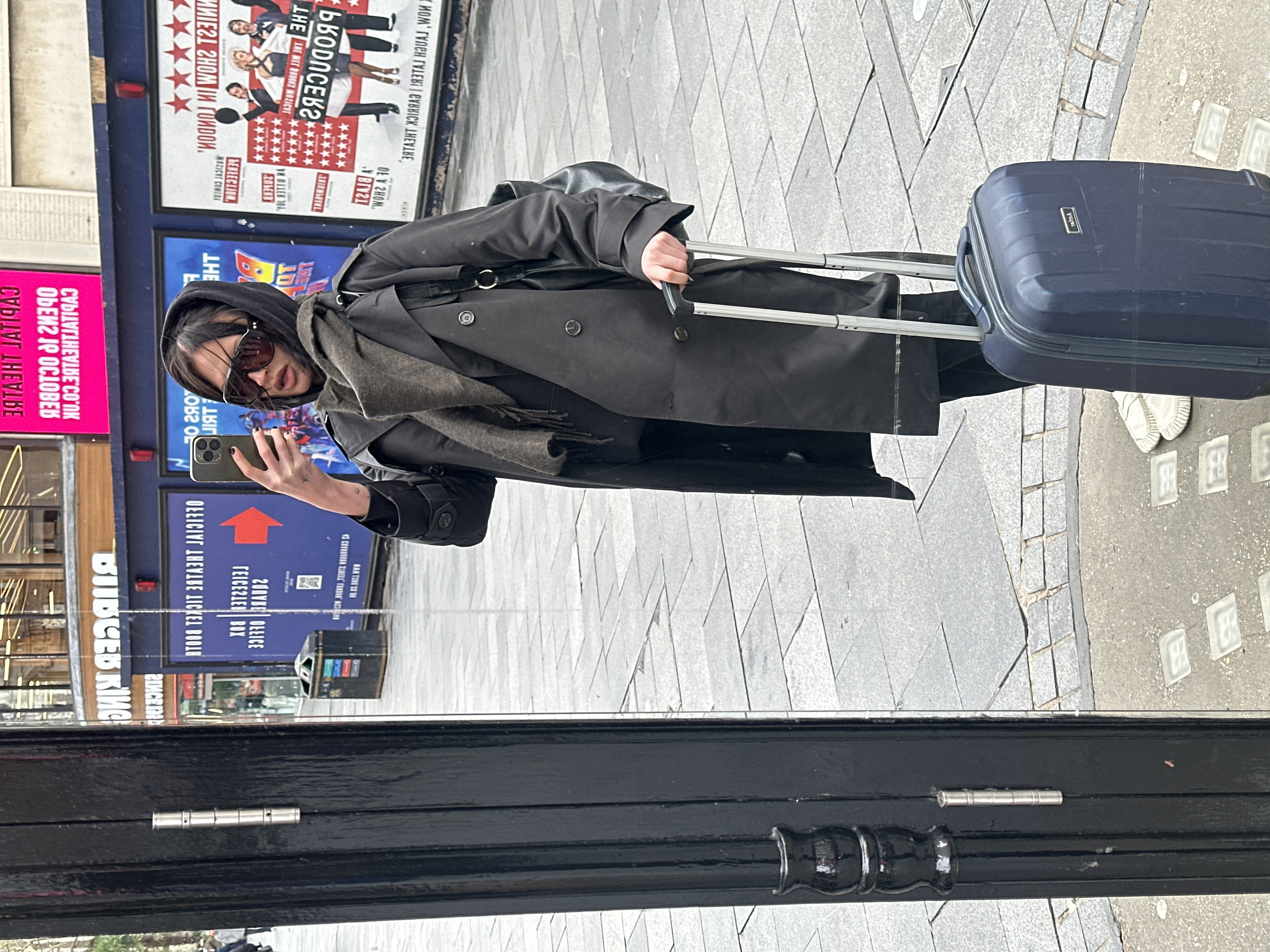 A single individual, appearing female, is depicted taking a selfie in a reflective black pillar on a paved street in Leicester Square, London, United Kingdom. The individual wears a dark trench coat, a dark hooded garment, a dark scarf, dark trousers, and light-colored slip-on shoes. Dark sunglasses obscure their eyes. They hold a dark smartphone with a triple-lens camera array in their right hand, positioned to capture their reflection. A dark backpack is visible on their left shoulder. Their left hand grips the extended handle of a dark blue rolling suitcase resting on the grey flagstone pavement. In the background, reversed reflections of urban street elements are visible, including a large blue billboard advertising "Official Theatre Ticket Booth Leicester Sq" and displaying posters for "The Producers" musical. Another pink sign, also reflected and reversed, reads "Capital Theatre." Glass paving lights are embedded in the pavement near the suitcase. A dark public waste bin is partially visible in the distant background.