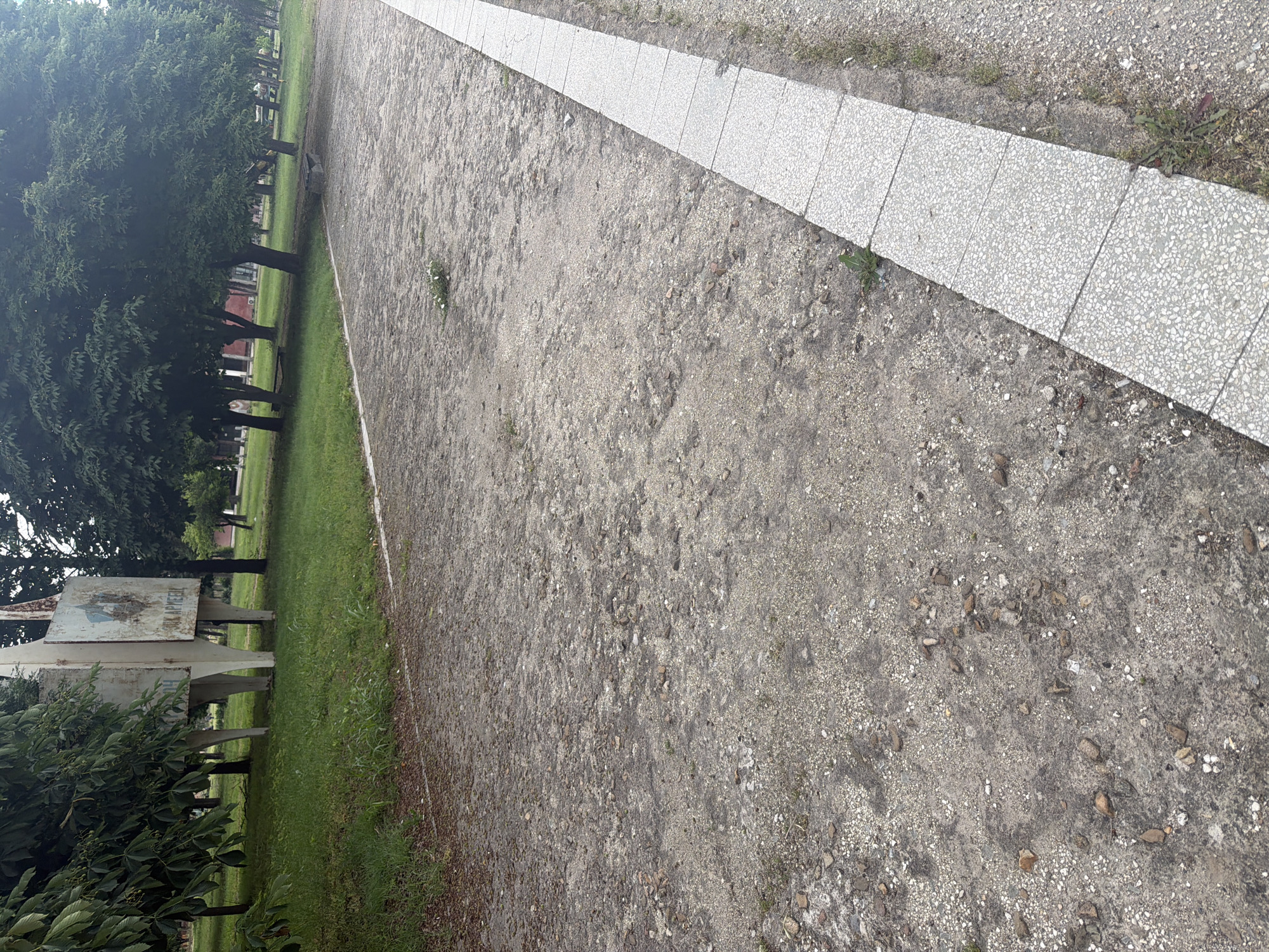 The image shows a path in Dolna Mitropolia, Bulgaria. The path is made of packed gravel and dirt, with a border of light gray, tiled pavers on one side. Along the other side of the path is a grassy area with trees and foliage. A sign is visible among the trees. The lighting suggests it is daytime.