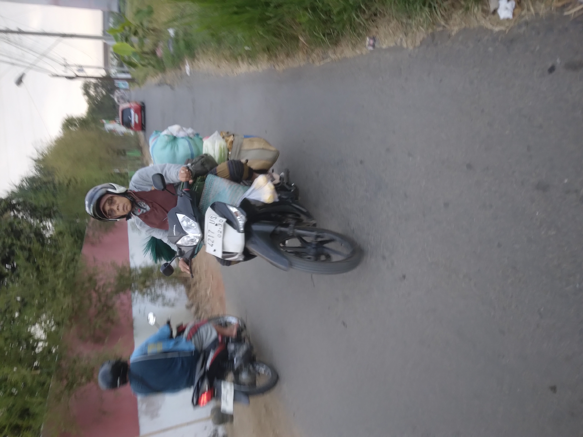 The image depicts a road scene in Desa Pamijen, Kecamatan Sokaraja, Indonesia. In the foreground, a person wearing a helmet and a maroon apron is riding a black motorcycle. The motorcycle is carrying several bags and bundles, including a large light blue bag and a tan bag behind the rider. The license plate of the motorcycle is visible and reads "4217 US 02.30". In the middle ground, another person is riding a red and black motorcycle, also wearing a helmet. This motorcycle also appears to be carrying items. Further down the road, a red car is visible. The environment includes trees and vegetation along the side of the road, as well as utility poles and wires. The road surface is asphalt. The sky is overcast.