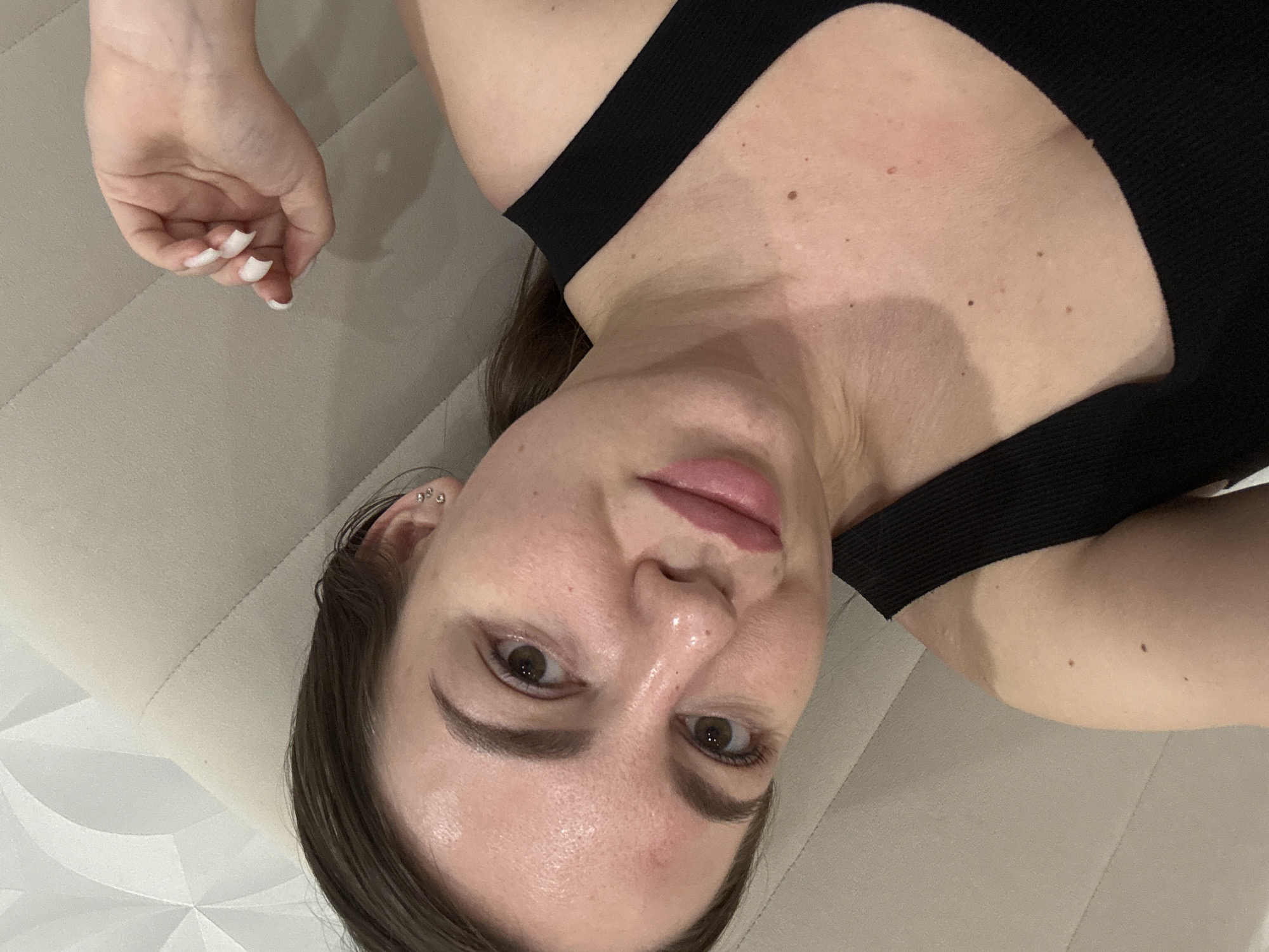 A woman is lying down on a light-colored surface, possibly a sofa or bed. She is wearing a black top with straps. Her head is positioned in the lower center of the frame, with her face looking directly at the camera. She has light skin, brown hair and eyes, and is wearing what appears to be neutral makeup. Her left hand is visible in the upper left corner of the frame with white nail polish. The photo is a close-up shot, with a plain background. There is no indication of the location being Orekhovo-Zuyevo, Russia.