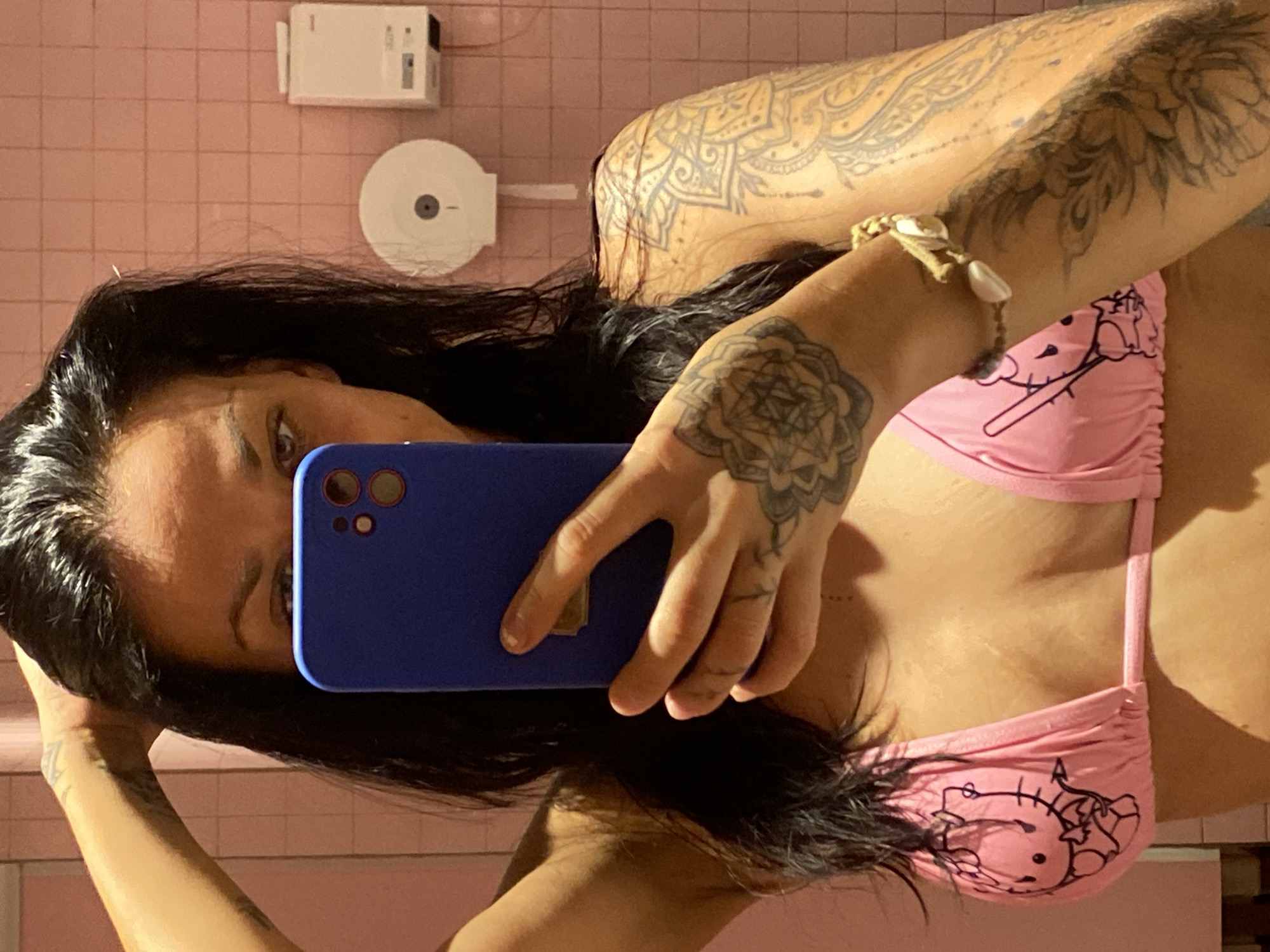 An individual is captured in a mirror selfie, with the image rotated 90 degrees clockwise from a typical upright orientation. The person, who appears to have long, dark hair, looks directly at their blue smartphone, which is held up to obscure part of their face. Their right eye and parts of their nose and mouth are visible.
The person is wearing a pink bikini top featuring a black line art design of what appears to be Hello Kitty. Intricate tattoos are visible on both arms and shoulders, including a large mandala-style pattern on the upper arm and shoulder, and another floral or geometric design on the forearm. A light-colored bracelet adorned with small shells or beads is worn on the right wrist.
The background consists of a pink tiled wall, suggesting an indoor setting like a restroom or changing room. Two white wall-mounted fixtures are visible: one, higher up, resembles an alarm sensor or electrical box, while the other, below it, is round and could be a toilet paper dispenser. The overall lighting is artificial and warm. The activity is clearly a mirror selfie being taken in Tien Thanh Ward, Vietnam. No discernible text is visible other than the implied "Hello Kitty" design.
