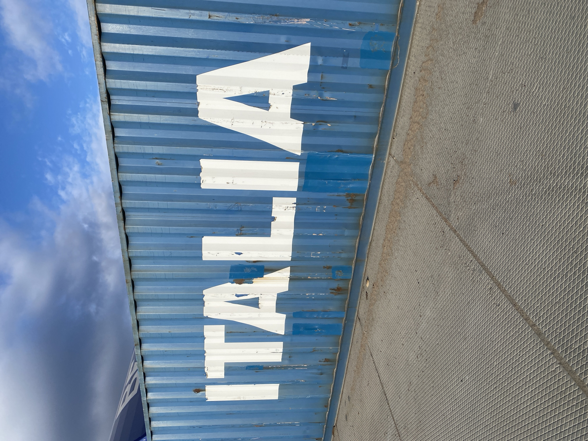 The image captures a slightly tilted, close-up view of a weathered blue shipping container in Luzhitsy, Russia. The container's corrugated metal surface is painted a faded blue, with significant chipping and rust visible in various spots, indicating wear and tear. Large, distressed white letters are stenciled vertically across the container, spelling "TALIA".
To the left and above the container, a bright blue sky with scattered white and grey clouds is visible, suggesting it is daytime. On the right side, a textured grey surface, resembling a metal grate or industrial flooring, runs parallel to the container. The overall setting appears to be an industrial area, likely a port or shipping yard, due to the presence of the container and the type of adjacent structure. No people or specific activities are depicted.