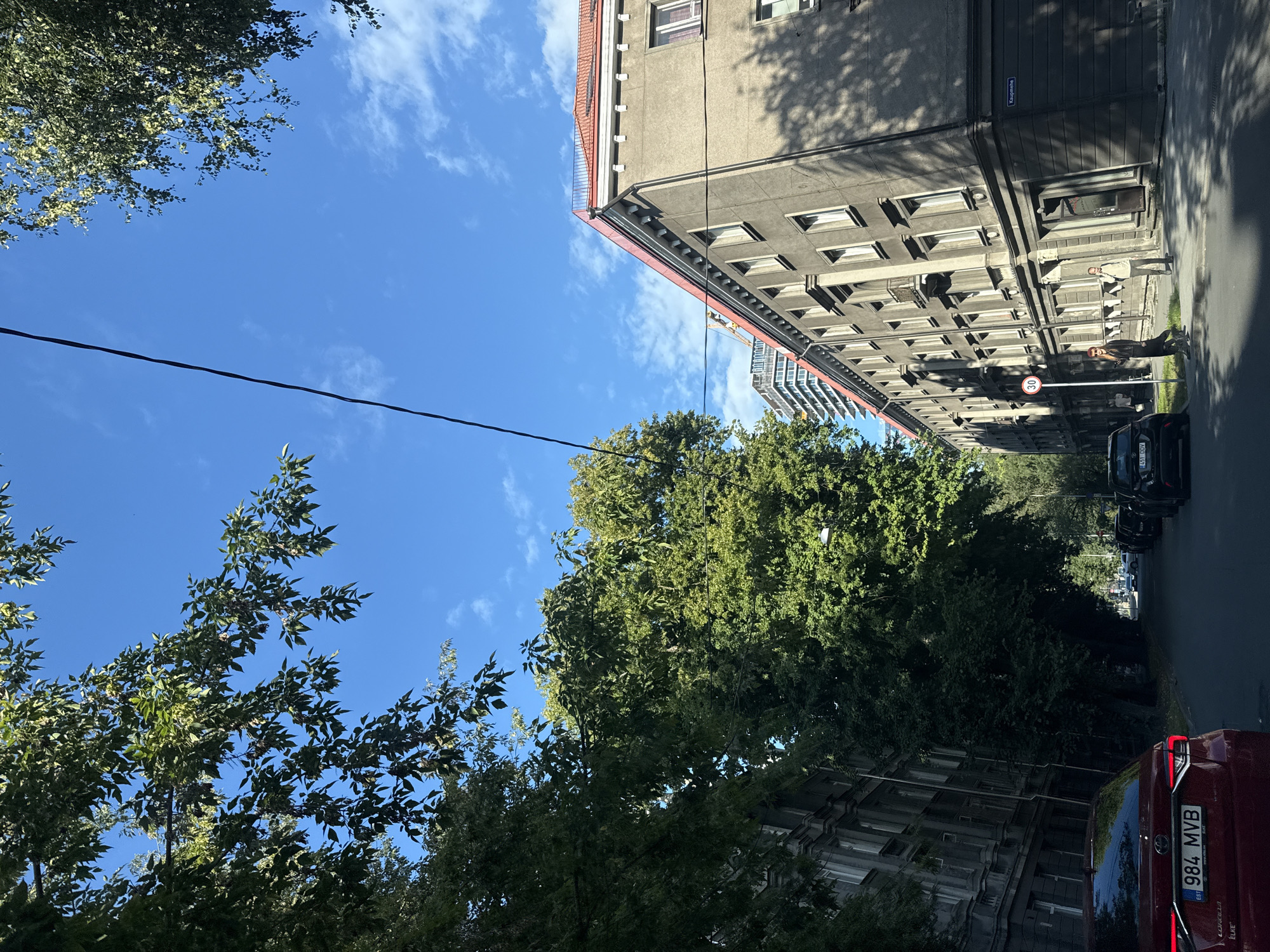 The image depicts a street scene in Tallinn, Estonia, on a clear day with a bright blue sky and scattered white clouds. Lush green trees line the left side of the street, partially obscuring a grey, multi-story building with numerous windows. On the right side of the street, a grey, older apartment building with a red-tiled roof dominates the view. This building has a row of windows on its upper floors and ground-floor entrances. A street sign reading "Kaugmehe" is visible on the facade of this building.
In the foreground, the rear of a red car with the license plate "984 MVB" is visible. Further down the street, several other cars are parked along the right side. Two individuals are walking on the sidewalk in front of the grey apartment building on the right. One person, wearing dark clothing and sunglasses, is walking towards the left, holding a phone. The other person, in lighter clothing, is walking towards the right, also appearing to be holding a phone. A circular speed limit sign indicating "30" is mounted on a pole near them.
In the background, a modern, tall building with a glass facade is visible behind the older apartment building. A construction crane is also present in the distance. The overall impression is of an urban environment with a mix of older and newer architecture, softened by the presence of mature trees.