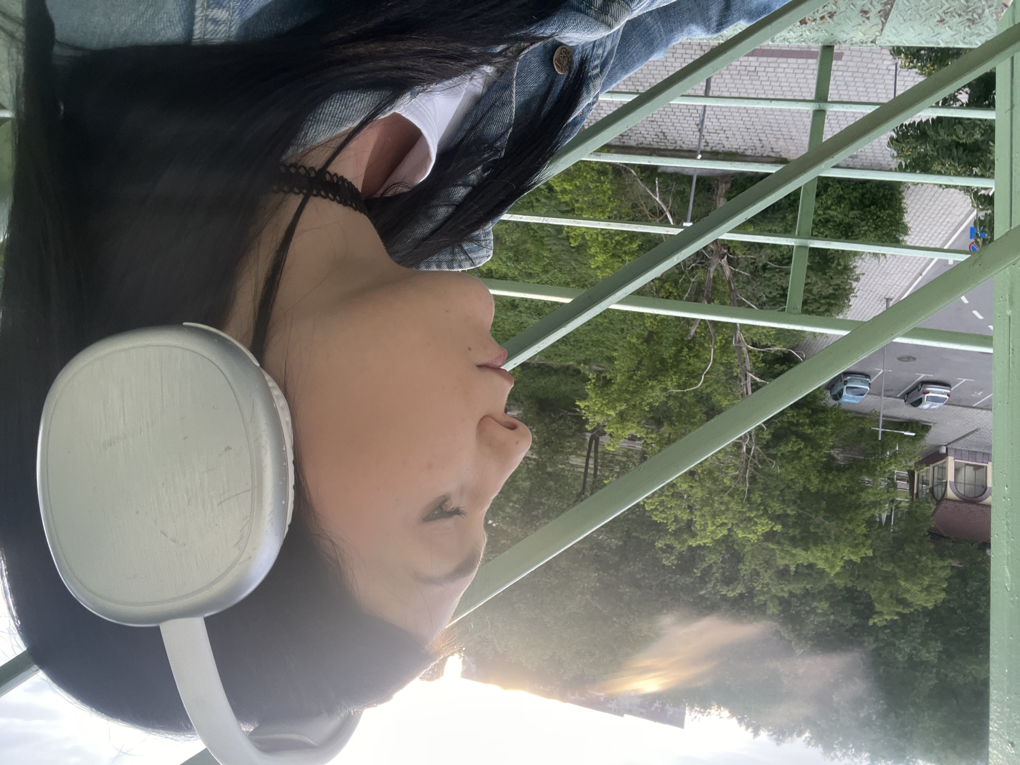 An overhead, outdoor portrait captures a person from the chest up, positioned on an elevated green metal structure, possibly a bridge or walkway. The image is rotated 90 degrees counter-clockwise, showing the person's right side in profile.
The subject is a young person with long, dark, straight hair flowing down their back. They are wearing large, silver or light gray over-ear headphones, which show some minor scuff marks. Around their neck is a black lace choker. They are dressed in a blue denim jacket with a metal button visible on the collar, over a white top. Their face is serene, with their lips slightly parted, and their gaze directed outwards and slightly upwards towards the horizon.
The green metal structure they are on features a grid-like pattern of beams and supports. Below and behind the person, the scene unfolds with lush green trees dominating the midground. Beyond the trees, paved areas, possibly a sidewalk or road with a white brick pattern, and a parking lot are visible, where at least two cars are parked. Further in the distance, parts of buildings are discernible. The lighting suggests late afternoon or early evening, with a soft, warm glow from the setting sun visible on the horizon, casting a hazy light through the air. The sky appears partially cloudy or hazy. No visible text is present in the image.