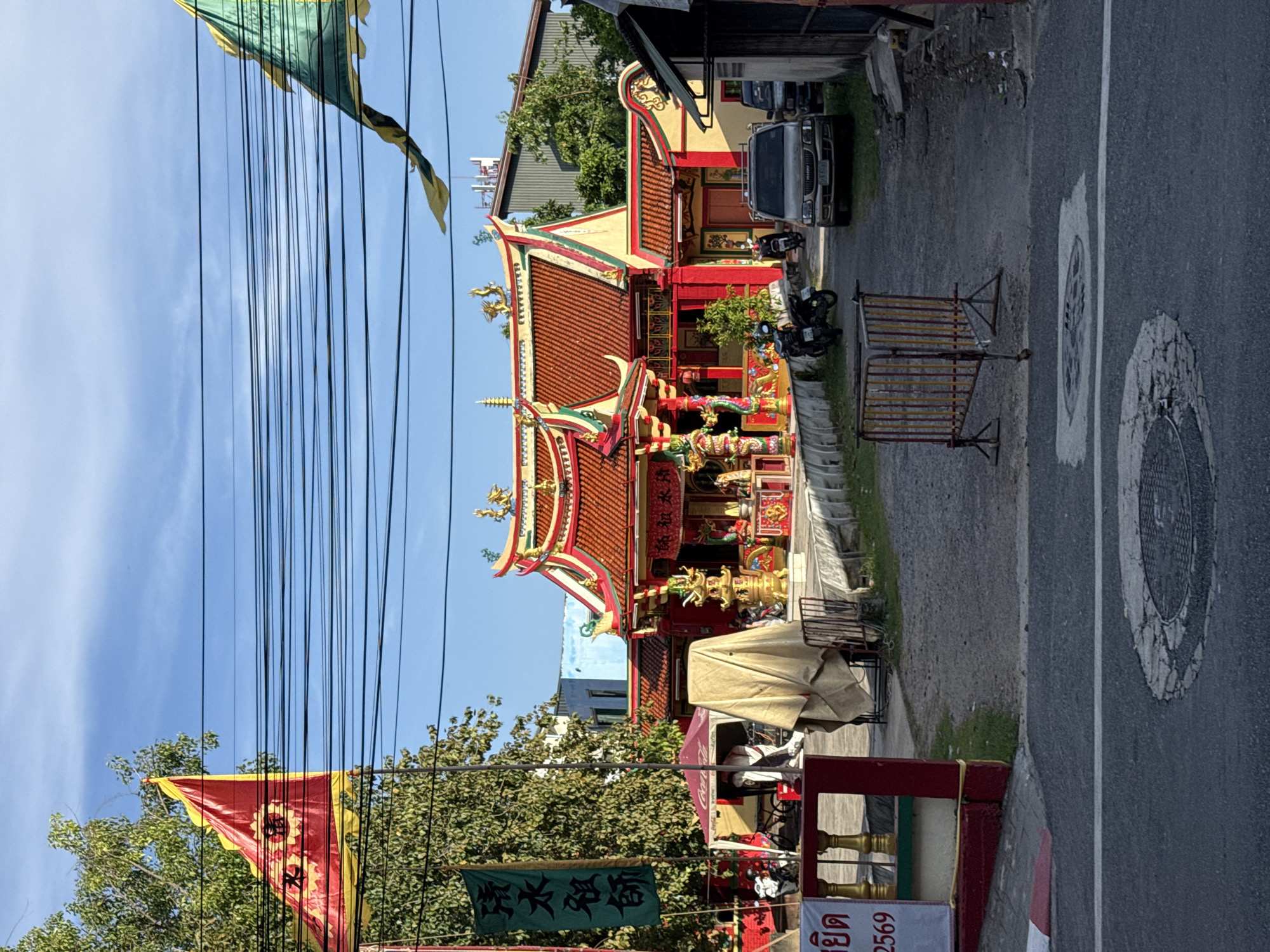 The image captures a frontal view of a traditional Chinese temple located in Phuket City Municipality, Thailand. The temple, adorned with vibrant red and gold colors, features an ornate roof with decorative sculptures. In the foreground, a street with a manhole cover is visible, with a temporary metal barrier placed on the side. Various elements contribute to the scene's cultural context, including Chinese flags and banners with inscriptions. Several motorcycles and a silver SUV are parked in the vicinity of the temple. The environment appears to be an urban setting with power lines crisscrossing the sky, indicating a developed area. The overall atmosphere is serene, with clear blue skies overhead.