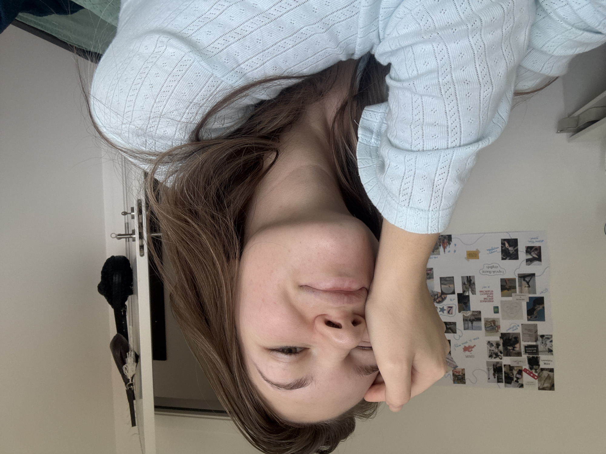 A woman is photographed from an upside-down perspective. She is wearing a light blue, patterned top and has brown hair. Her face is visible with her eyes slightly closed, and one hand is touching her face near her eye. In the background, there is a door with a black object hanging from it, a white wall, and a collage of pictures adhered to the wall. The location is not discernible from the given image.