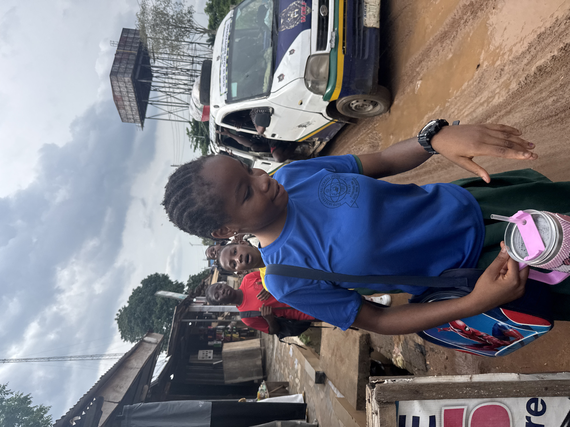 The image captures a bustling street scene in Ihiagwa, Nigeria, under an overcast sky. The foreground is dominated by a young girl, possibly a student, walking along what appears to be a muddy, unpaved road. She wears a bright blue short-sleeved t-shirt with a school crest visible on her left chest, and a dark skirt. The circular crest reads "INTERNATIONAL SCHOOL (FISO)" and "FOTO INTERNATIONAL" with the motto "TO EXCEL AND LEAD". On her left wrist, she wears a black digital watch. She carries a shoulder bag featuring a Spider-Man design and holds a pink-lidded cup with a straw in her right hand. Her expression is neutral.
Behind her, other people are visible, including another young person in a blue shirt and a figure in a red t-shirt further back, near makeshift market stalls with corrugated metal roofs. In the mid-ground, a white and blue minibus, characteristic of local commercial transport, is parked on the muddy road. Text "WOLF" is visible on the front of the bus, along with "ABIA" and what appears to be a license plate number "ABJ-704". Faded text resembling "GOD IS GOOD" can also be seen on its side. Inside the bus, a person's head is visible.
In the background, a large, elevated rectangular water tank or tower stands against the cloudy sky, hinting at public infrastructure. The ground is consistently muddy, suggesting either recent rain or poor road conditions. The overall atmosphere depicts an everyday street scene in a semi-urban or rural Nigerian setting.