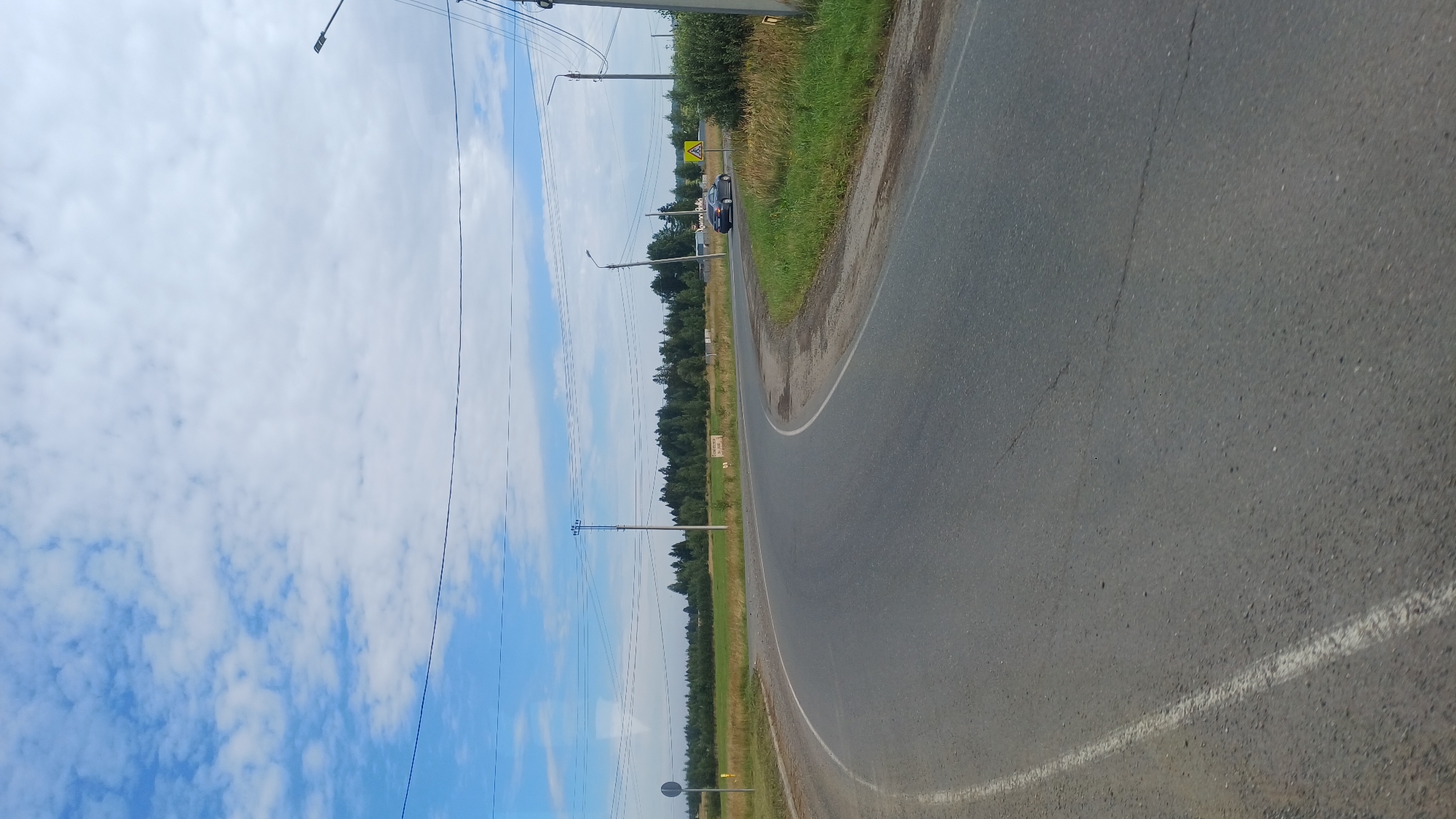 A paved road curves to the right in a rural setting. The road has a white line on its left edge. To the left of the road, there is a grassy verge. Further left, there is a field with some trees in the distance. To the right of the road, there is a grassy area with tall grass and some bushes. In the distance, there is a line of trees. A few power lines are visible overhead, supported by poles. There are two streetlights visible on the right side of the road, and a yellow pedestrian crossing sign. A dark-colored car is driving on the road in the distance, on the right side. The sky is blue with scattered white clouds. The image appears to be taken in the daytime. The location is identified as Kamennoye, Russia.