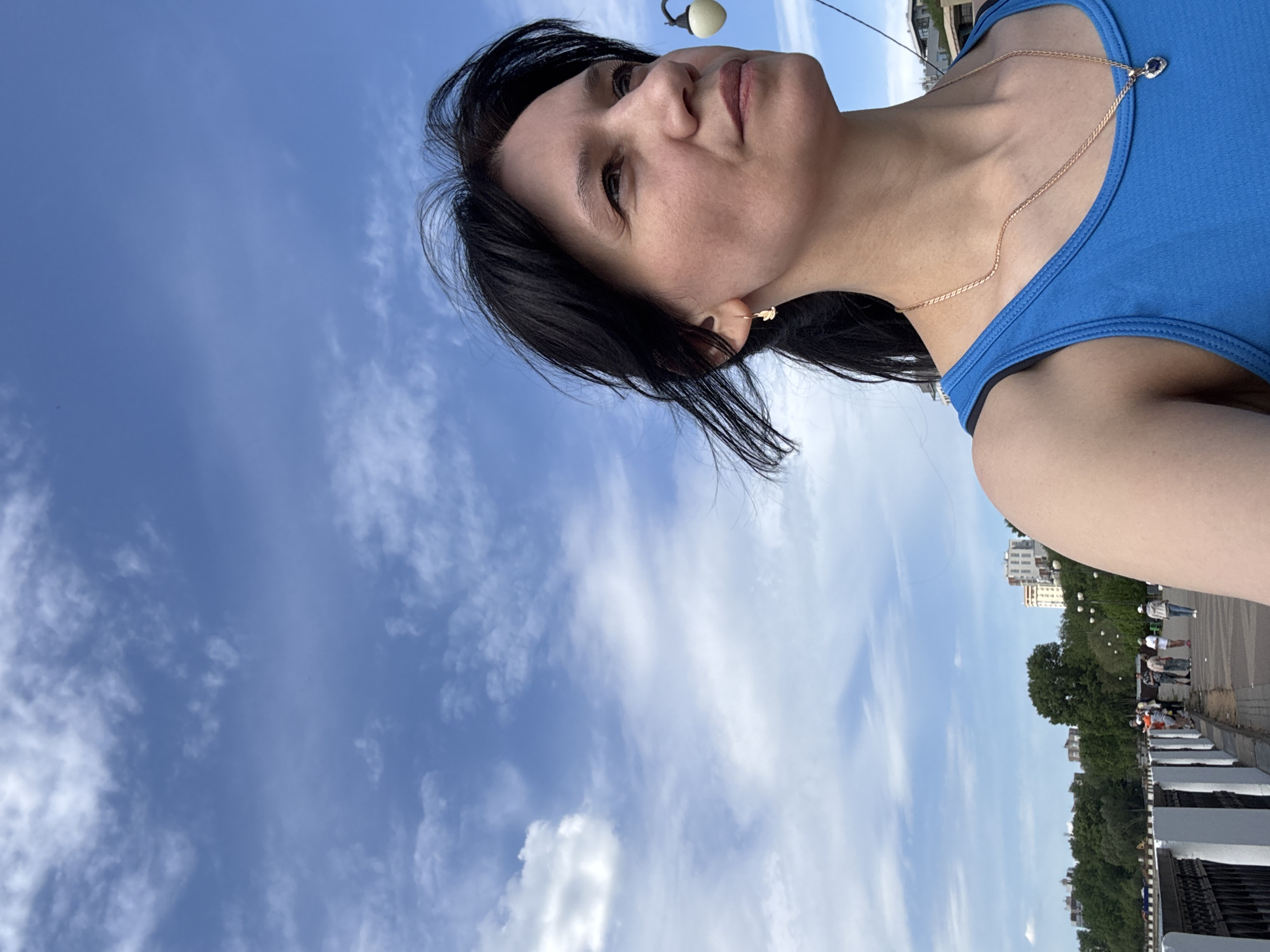 A photograph shows a woman with dark hair wearing a blue tank top and a gold necklace, oriented with her head towards the upper left. She has a gold earring in her left ear. The background features a blue sky with scattered clouds. In the distance, a park area with trees and buildings can be seen. A streetlight is visible in the upper center of the frame. The location appears to be a city sidewalk or bridge.