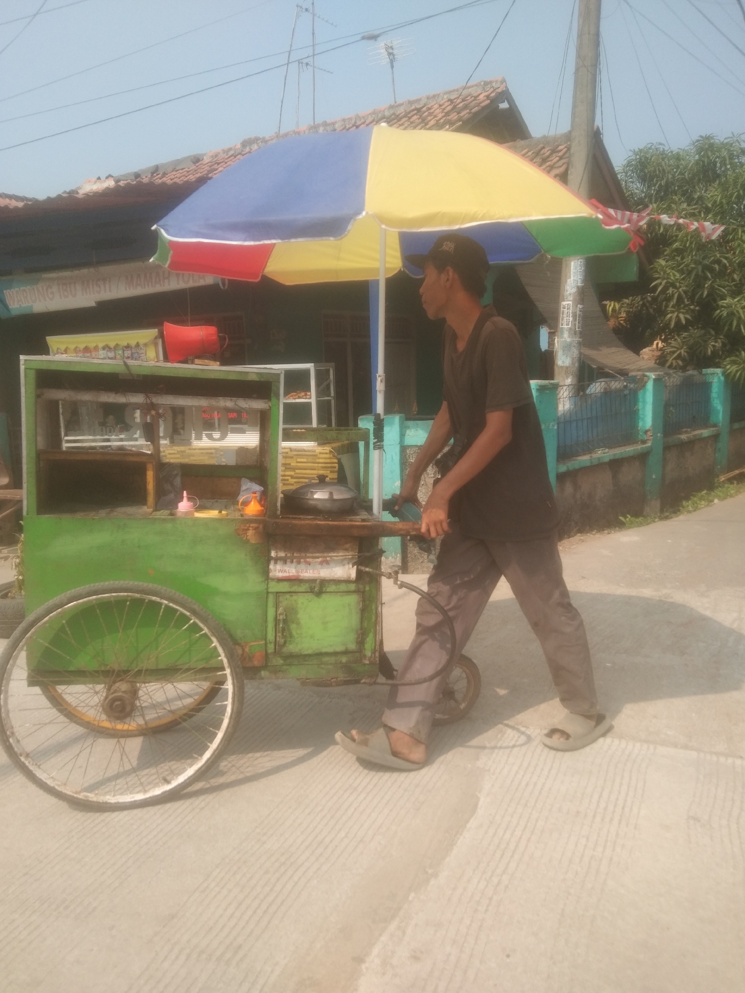 A man pushes a green food cart with a colorful umbrella on a sunny day in Subang, Indonesia. The cart is adorned with text that is partially legible and appears to read "WARUNG IBU MISTI / MAMAH YULA". The man is wearing a dark t-shirt, grey pants, and sandals, and he is pushing the cart by its handle. Behind the cart, a building with a tiled roof and a light blue fence can be seen. The ground is a paved road with a linear pattern. The scene is outdoors, with a clear blue sky.