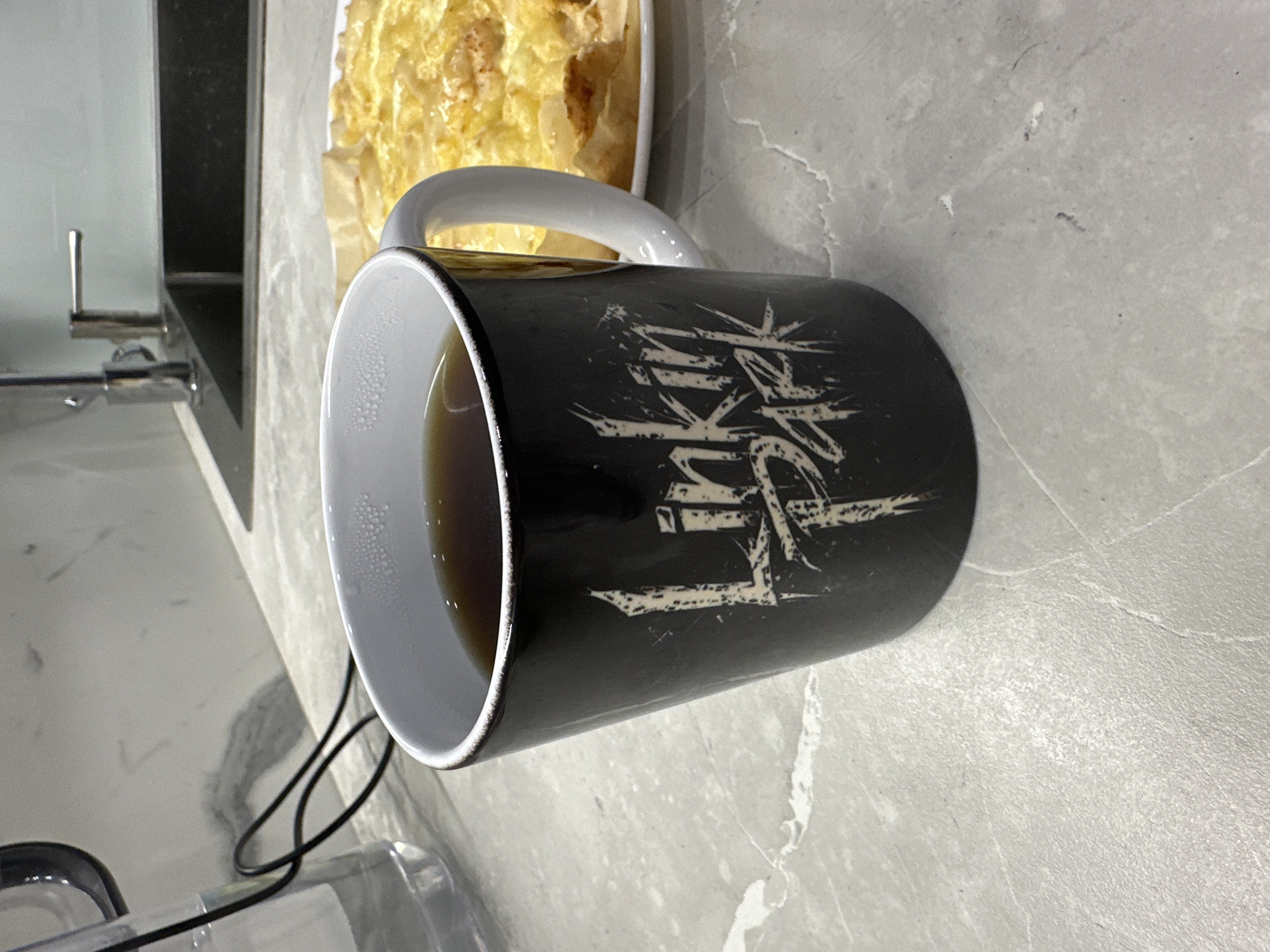 A high-angle medium shot shows a black mug with the band "Linkin Park" logo on it, filled with a dark liquid, resting on a grey countertop. To the right of the mug is a plate of what appears to be quiche or tart. In the background, a kitchen sink and faucet are visible, along with a clear plastic pitcher on the left. The scene is indoors, in what seems to be a domestic kitchen environment. The lighting is ambient, suggesting it may be daytime. There are no people visible in the image.