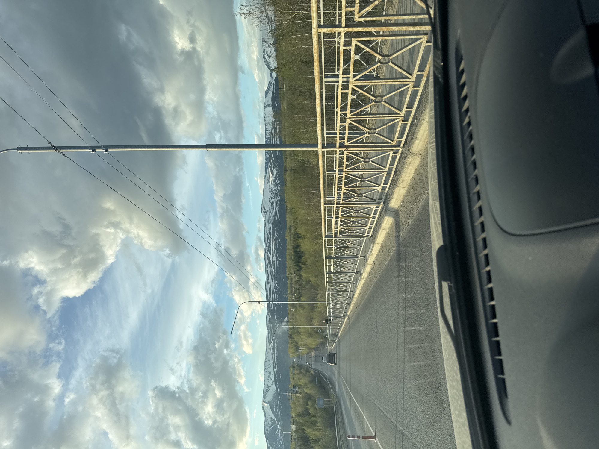 The image shows a view from inside a vehicle, likely a car, driving along a road or bridge in Apatity, Russia. The road has multiple lanes and is separated by a median. A metal guardrail runs along the side of the road. Streetlights line the road. In the distance, mountains covered with snow and vegetation are visible. The sky is partly cloudy with a mix of dark and light clouds. The dashboard of the car is visible on the right side of the image.