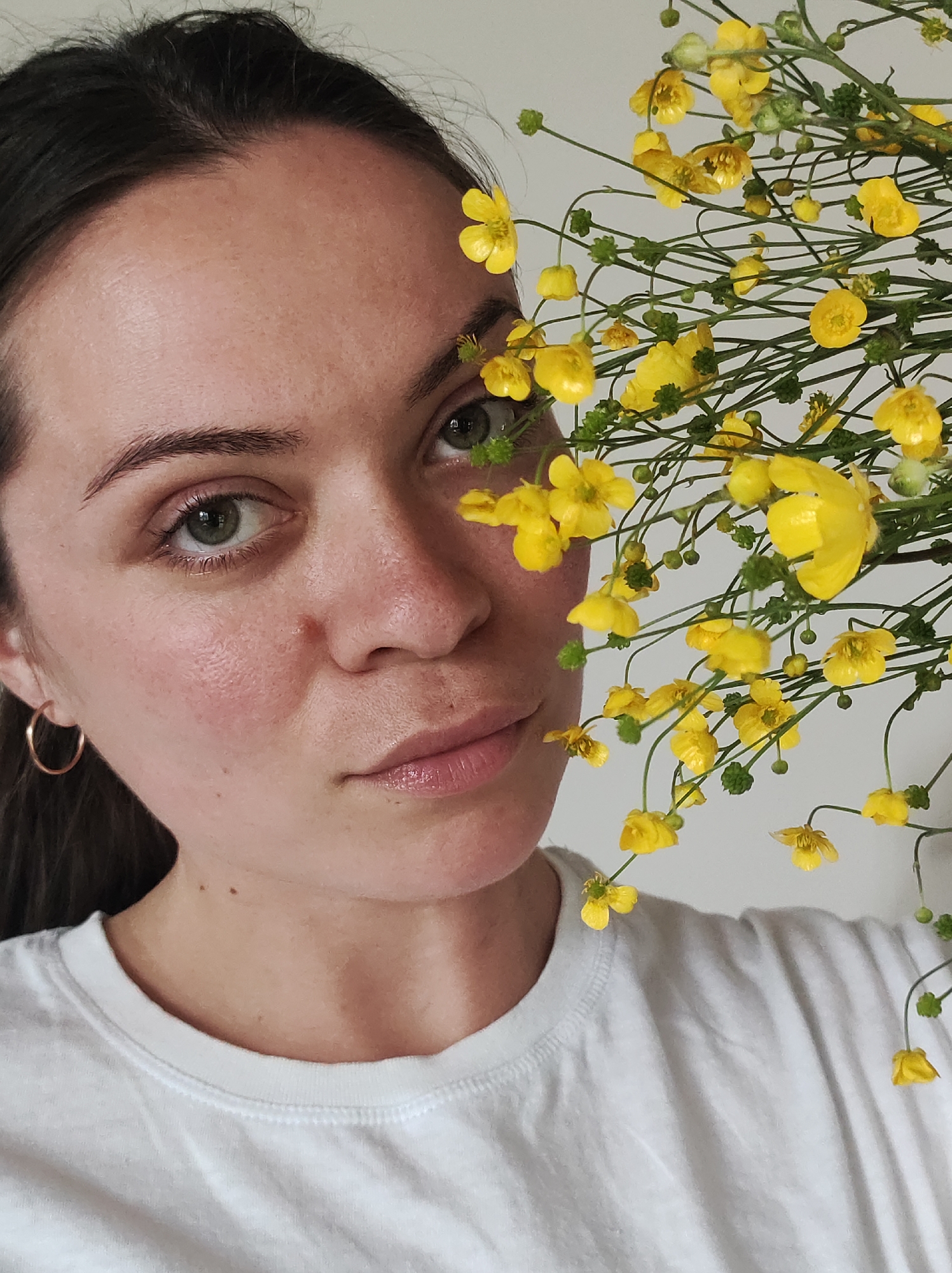 A close-up, eye-level portrait features a young woman looking directly at the viewer with a calm, neutral expression. Her face occupies the left side of the frame, while a dense cluster of vibrant yellow flowers, possibly buttercups, fills the right side, partially obscuring her and creating a natural frame.
The woman has fair skin, with a natural flush on her cheeks, and striking green eyes. Her dark brown hair is pulled back, with her hairline visible at the top. She has well-defined eyebrows and full, naturally colored lips with a slight sheen. A small, golden hoop earring is visible in her left ear. She is wearing a simple white crew-neck t-shirt.
The yellow flowers, with their delicate green stems and small buds, are in sharp focus, adding a bright contrast to the woman's features. The background is a plain, light-colored wall, indicating an indoor setting. The lighting is soft and even, highlighting the details of her skin and the texture of the flowers. The overall mood of the portrait is serene and intimate. There is no visible text in the image. The provided location context is Фоки, Russia.