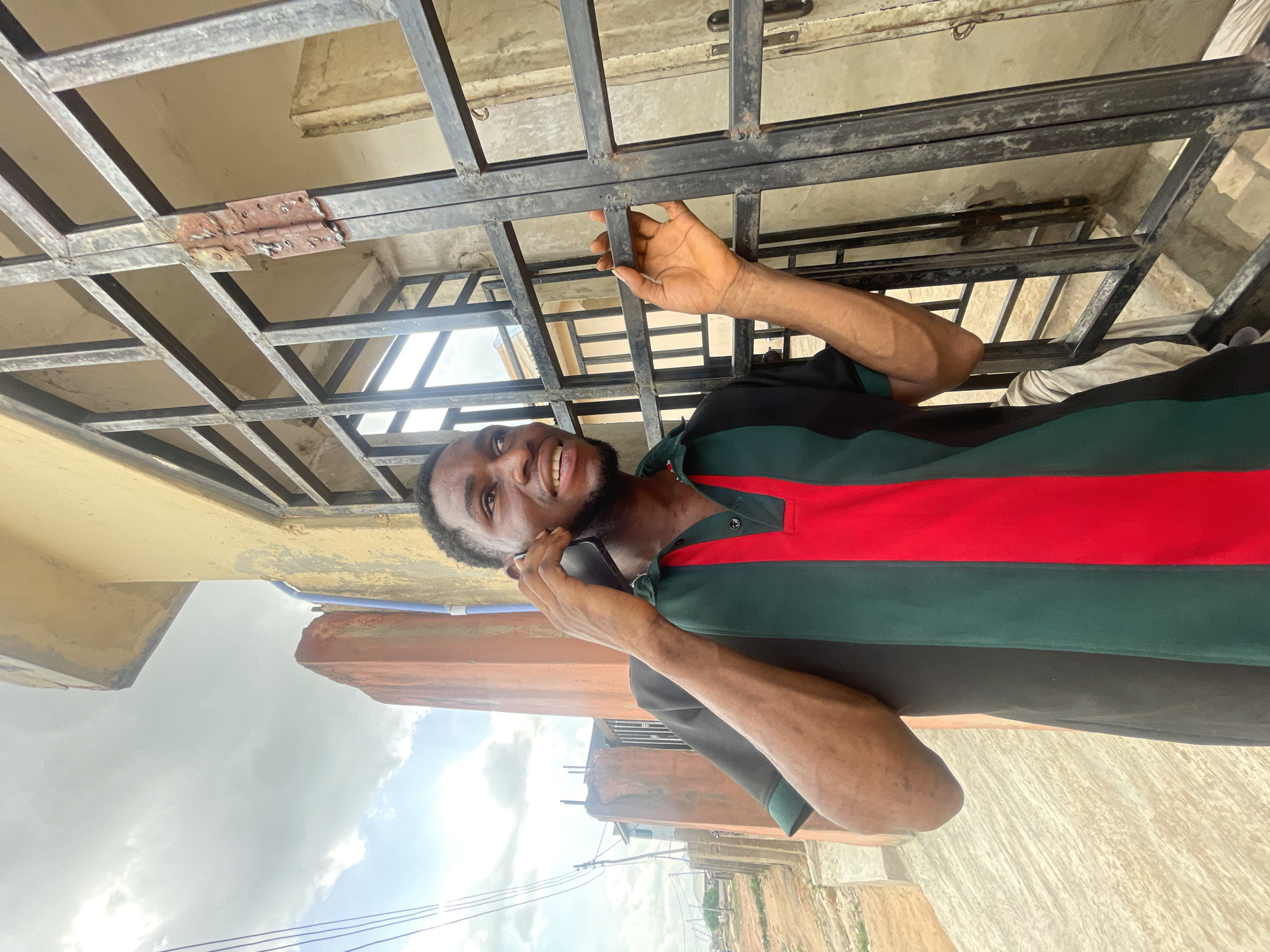 The image shows a man outdoors in Lokoja, Nigeria, apparently on a phone call. The man is dark-skinned, with a beard and short black hair. He is wearing a polo shirt with black shoulders, a green torso, and a horizontal red stripe. He holds a dark-colored smartphone to his ear with his left hand.
He stands beneath a dark-colored metal grating or framework, possibly part of a gate or window protection. Above the grating is a concrete structure, possibly part of a building. In the background is a cloudy sky and other buildings and possibly natural landscapes characteristic of Lokoja. The ground appears to be sandy or dusty. A blue pipe is visible near the man.