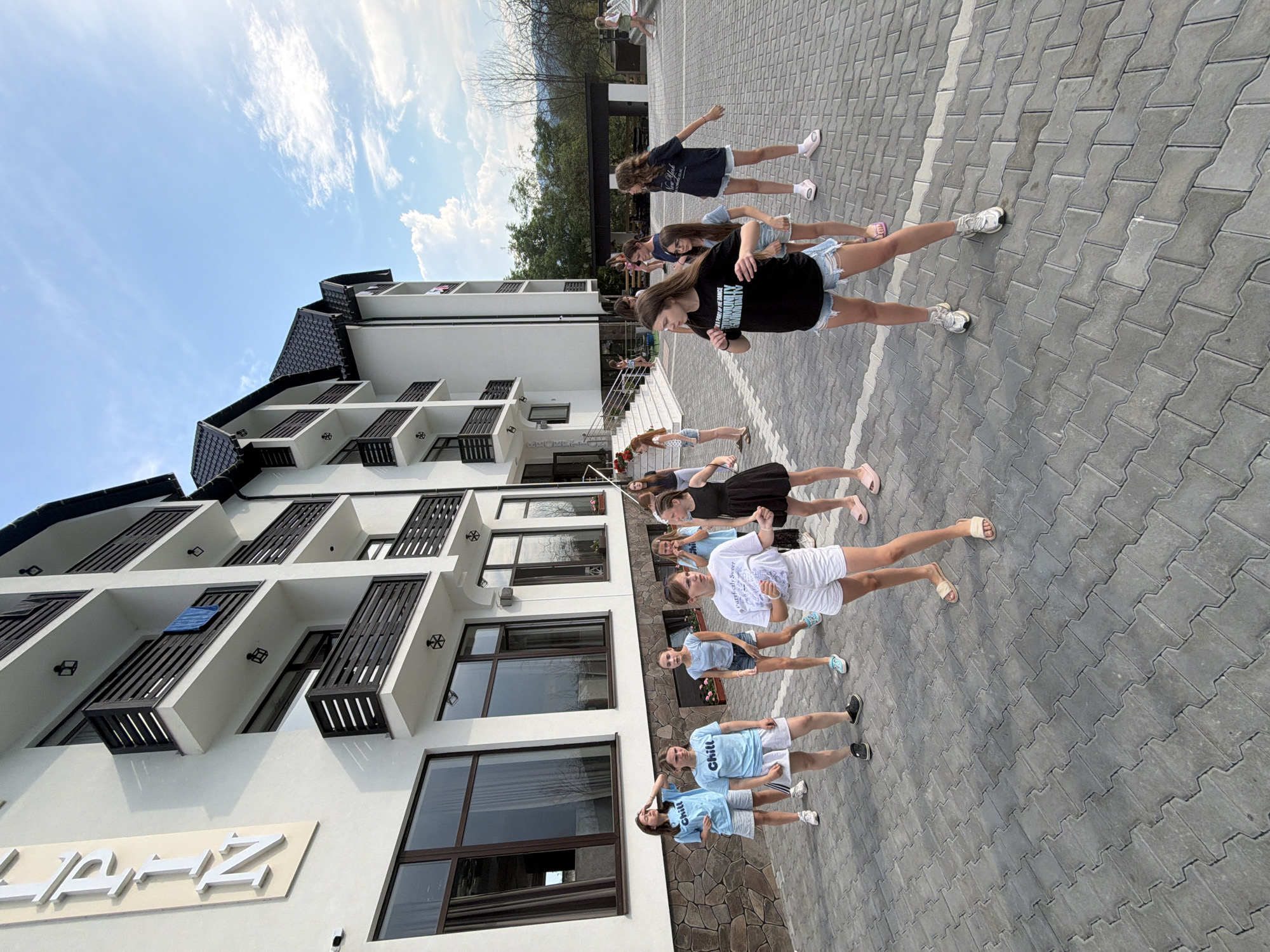 A group of young girls are standing in a line on a paved area in front of a modern white building with multiple balconies. The building appears to be a hotel or resort, with a sign visible at the bottom left that reads "LAZ". The girls are all dressed in casual summer clothing, including t-shirts and shorts. Some are wearing sneakers, while others are in sandals or flip-flops. They appear to be posing or participating in an activity, with some looking at the camera and others looking forward or to the side. The setting is outdoors, with a bright blue sky and white clouds visible in the background. Trees and a hillside can be seen behind the building. The ground is paved with interlocking grey stones. The time of day appears to be daytime, likely late morning or early afternoon, given the brightness of the sunlight. The overall mood seems relaxed and cheerful.