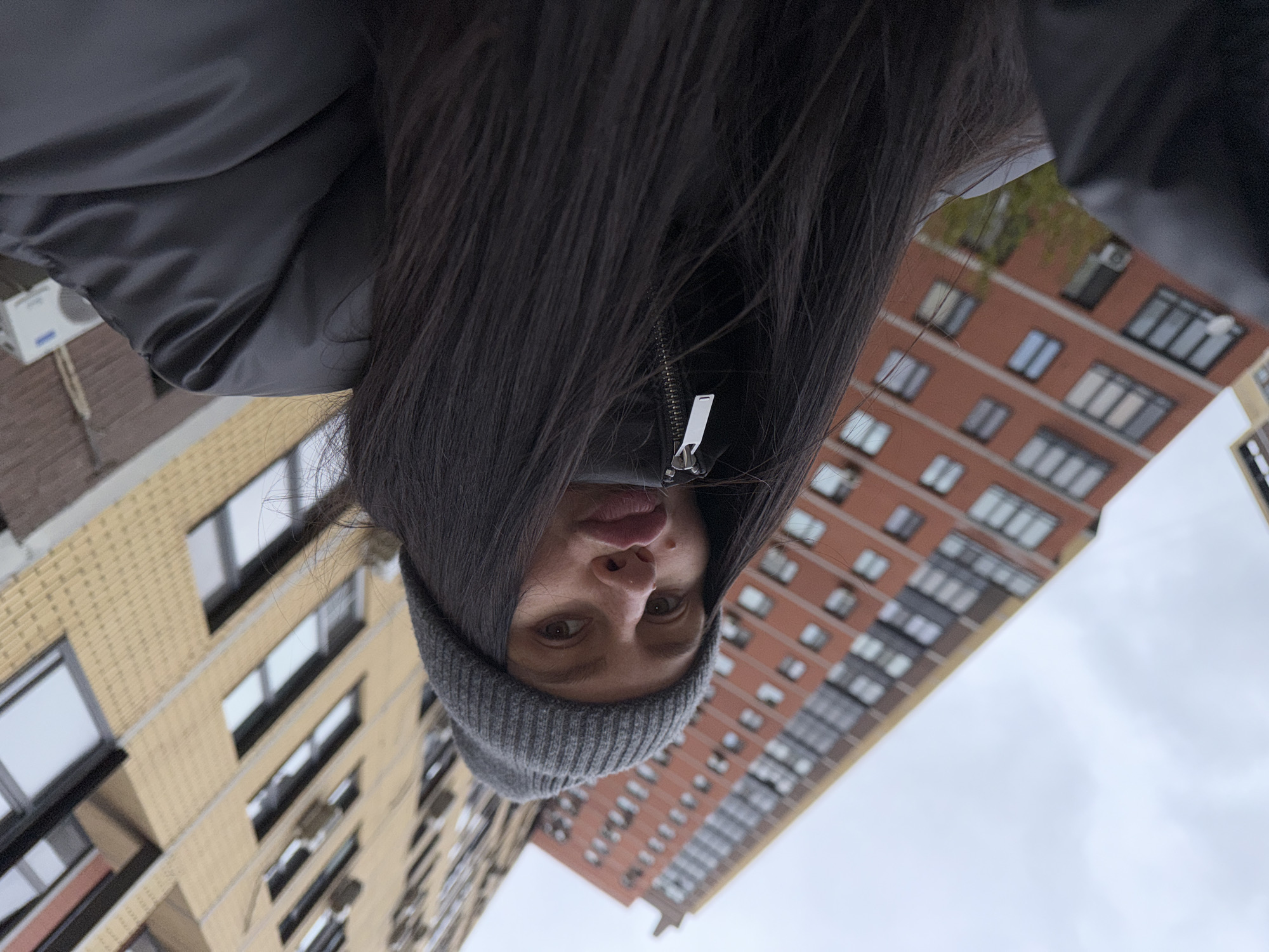 A selfie of a woman with long, dark hair wearing a gray beanie and a black jacket, possibly in Khimki, Russia, judging by the architecture in the background, including apartment buildings with distinct brick facades and window patterns. The photo is taken at a low angle, tilted, showing buildings and sky behind her. The sky is overcast.