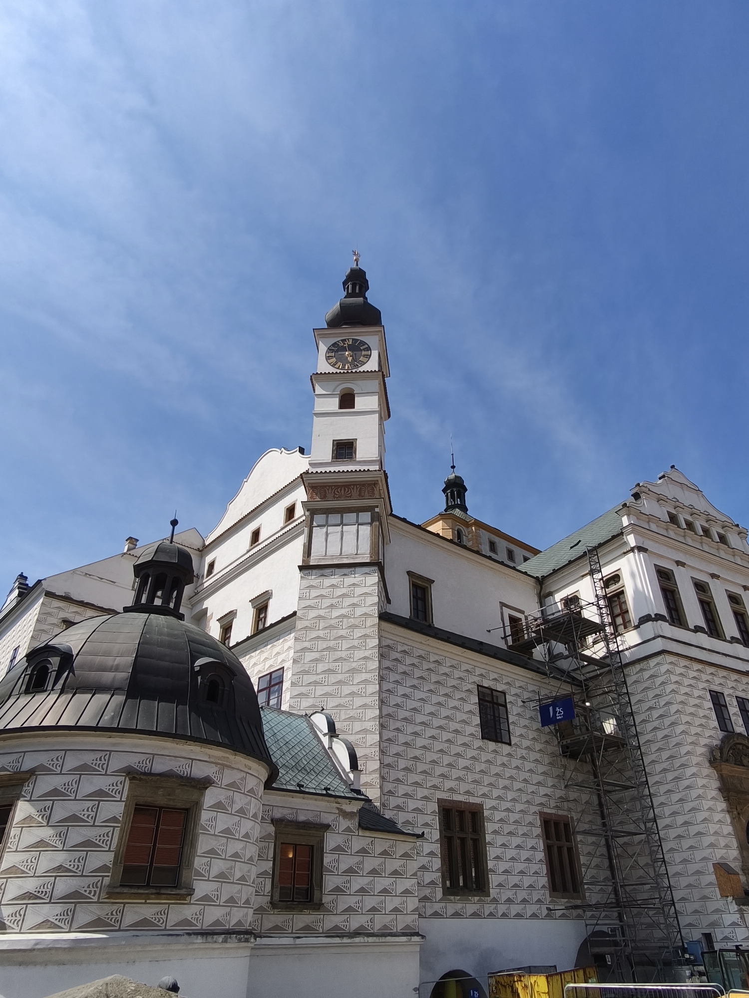 This is a medium-angle, upright shot of a historic building in Pardubice, Czechia, under a clear blue sky with sparse clouds. The building, primarily white, features a prominent clock tower with a black spire, adorned with Roman numerals on its clock face. The tower has decorative stone accents and several windows.
The main structure exhibits a unique sgraffito pattern of light grey triangular shapes against the white facade. To the left, a dark, domed structure with dormer windows is visible, also decorated with the sgraffito pattern. The building appears to be a palace or castle, with multiple wings and architectural elements, including a green-tiled roof.
Scaffolding is erected along one side of the building, suggesting renovation or maintenance work. A blue sign with white text "125" is attached to the scaffolding. Below the scaffolding, yellow construction bins are visible. The lighting suggests it is daytime, likely late morning or early afternoon, given the bright sunlight and clear skies. The overall scene conveys a sense of historical grandeur and ongoing preservation efforts.