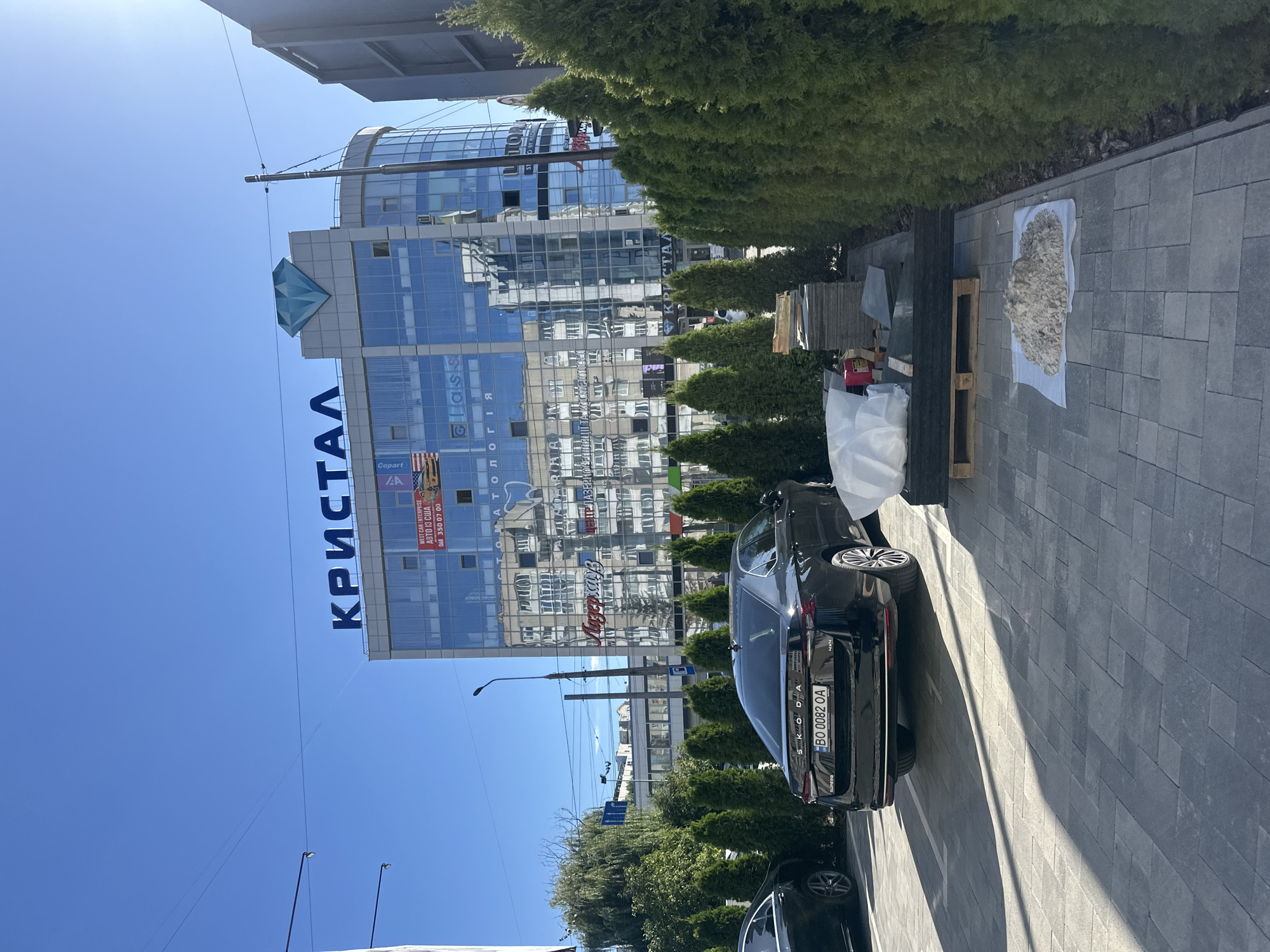 The image shows a frontal view of the "Kristal" building in Ternopil, Ukraine. The building is modern with a glass facade and a large blue diamond logo at the top. The ground floor appears to be a commercial establishment with various signs. In front of the building, there is a parking area with a black Skoda car and several cypress trees lining the perimeter. The sky is clear blue, indicating a sunny day. There is construction material on the ground near the trees, suggesting ongoing renovation or landscaping work.