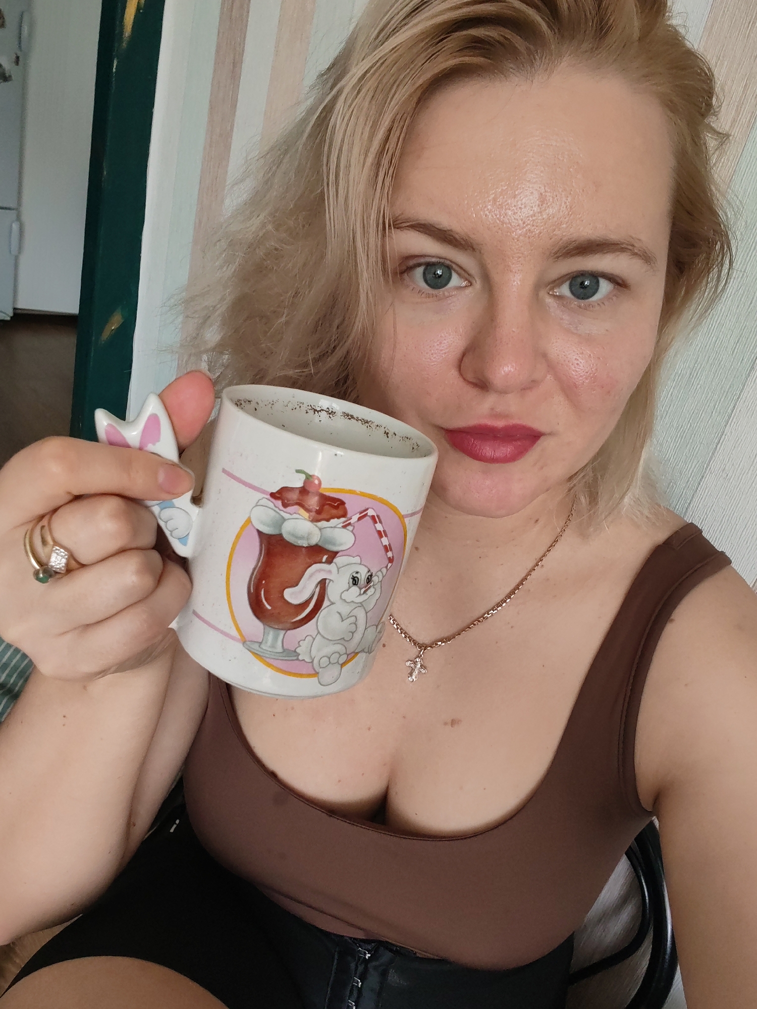 The image shows a close-up, selfie-style shot of a young Caucasian woman holding a mug. She has blonde hair, blue eyes, and is wearing red lipstick. She is wearing a brown tank top and a necklace with a cross pendant. The mug has a cartoon drawing of a bunny and a milkshake on it, with a bunny ear-shaped handle. The background appears to be a wall with striped wallpaper. The woman is indoors, likely in a residential setting, as suggested by the presence of a refrigerator in the background on the left side of the frame. There are no visible indicators that the image was taken in Izhevsk, Russia.