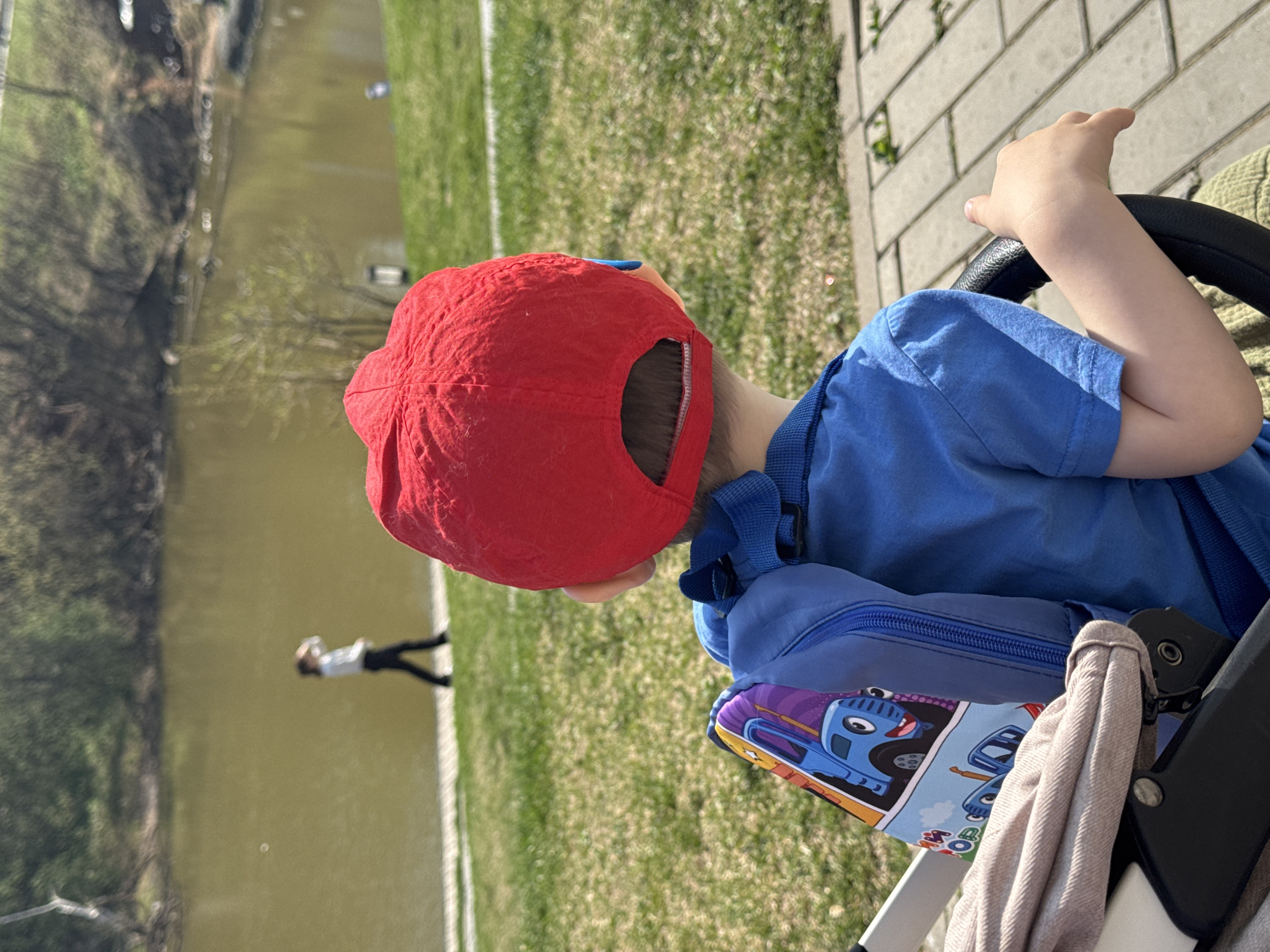A child wearing a red cap and blue shirt is sitting in a stroller facing away from the camera. They are near a body of water, likely a pond or canal, next to a grassy area and a brick walkway. A person is walking in the distance near the water. The stroller has a blue backpack attached with cartoon car designs. The location is Izhevsk, Russia.