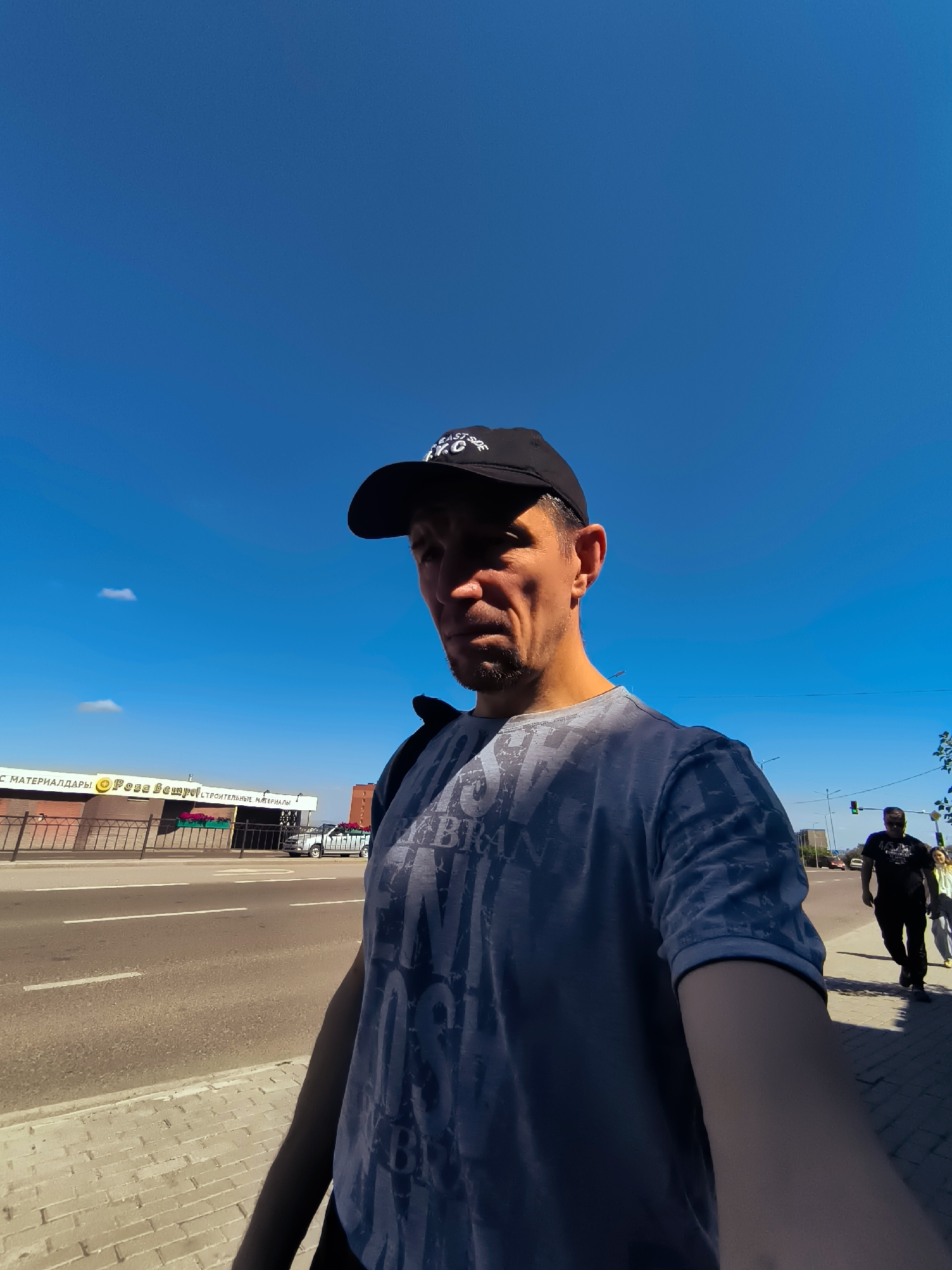 A man wearing a black baseball cap and a grey t-shirt is walking on a sidewalk in Qarağandı city administration, Kazakhstan. The man is facing the camera and is partially in shadow. In the background, there is a building with a sign that reads "РОЗА ВЕПРO" and "СТРОИТЕЛЬНЫЕ МАТЕРИАЛЫ". There are also other people walking on the sidewalk and cars driving on the road. The sky is blue and clear, with a few white clouds.
