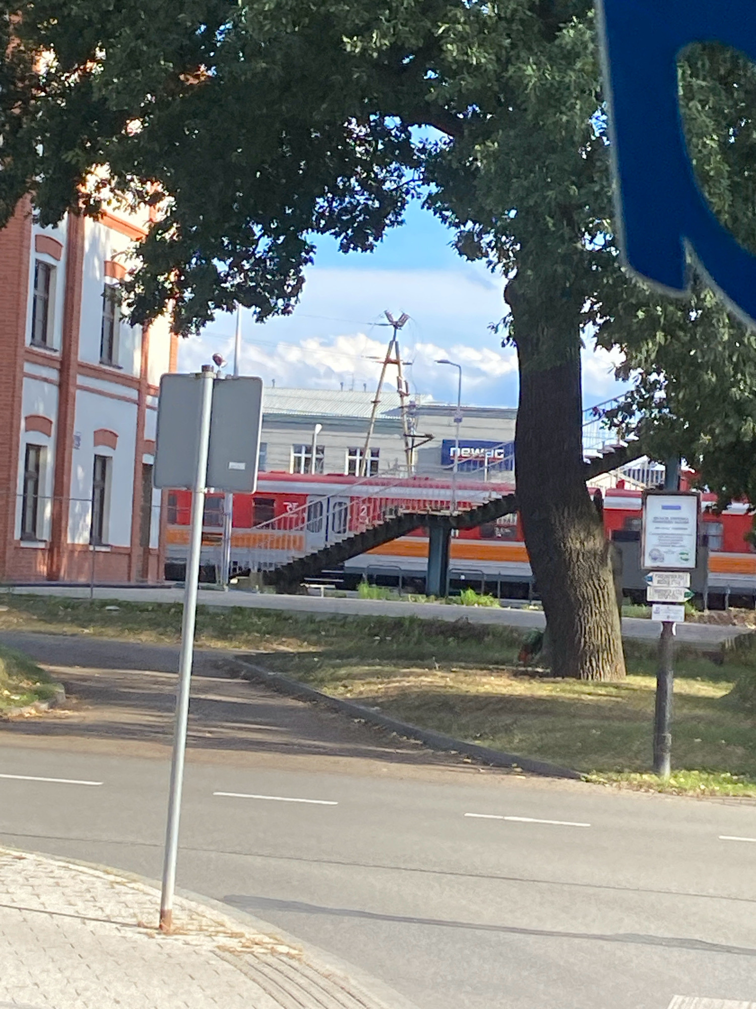 The image depicts a scene in Nowy Sącz, Poland, featuring a red and orange train at a station. To the left, a brick building with white trim and multiple windows is visible, partially obscured by a large tree with dark green foliage. In front of the building and tree, a silver pole with a gray sign is planted on a paved area. Behind the train, a modern building with "newag" signage is present, along with a pedestrian overpass with stairs. The train appears to be stationary. The environment suggests an outdoor, daytime setting with a clear sky.