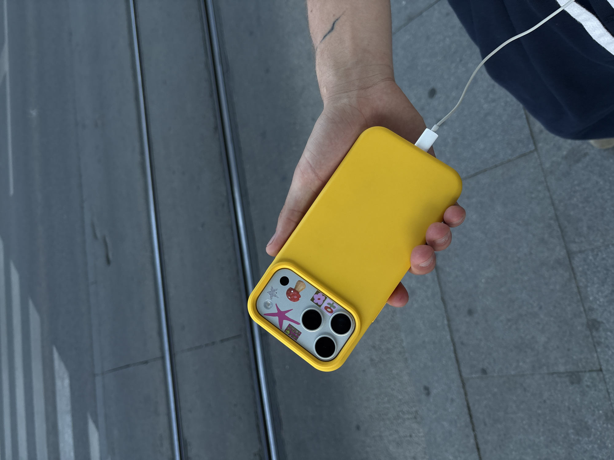 A person is holding a yellow smartphone in a matching yellow case. The phone is connected to a white charging cable. The back of the phone features several cartoon stickers. The person's hand is visible, and they are wearing dark clothing with white stripes. The background consists of a gray paved surface with tram tracks and a crosswalk. The photo was taken in Belgrade, Serbia.
