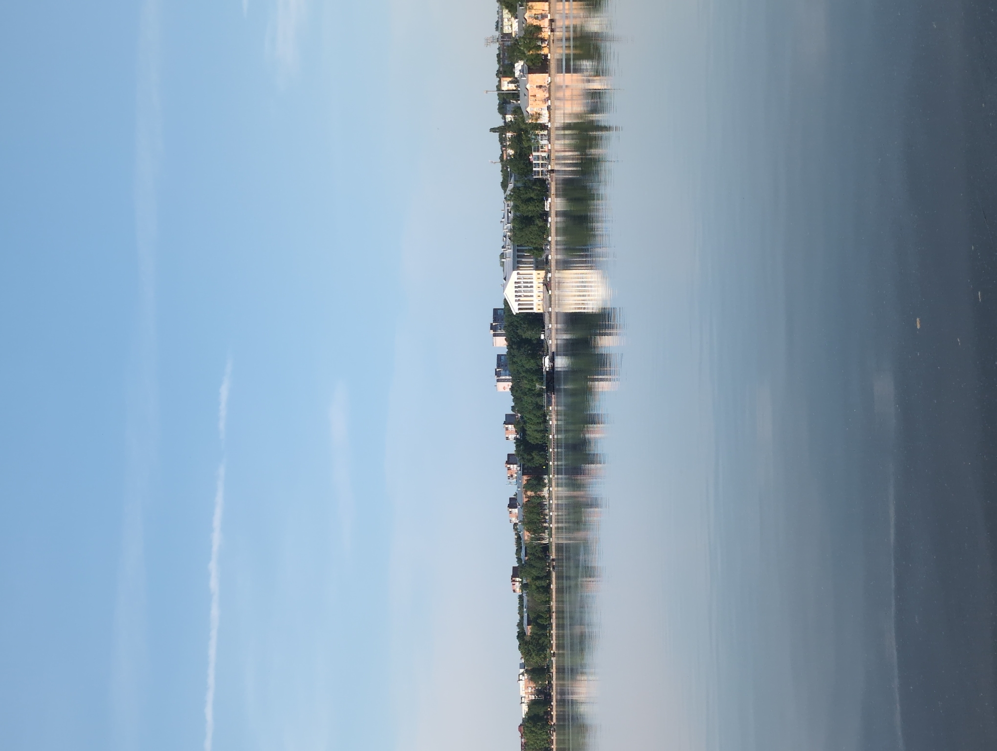 The image shows a waterfront scene in Votkinsk, Russia, with buildings reflected in the water. A bright blue sky is visible on the left, contrasted by the dark surface of the water on the right. The buildings, separated by trees, stretch along the waterfront. The reflections of the buildings and trees are visible in the water. The reflections distort and blur towards the center of the image. There are long narrow clouds in the blue sky.