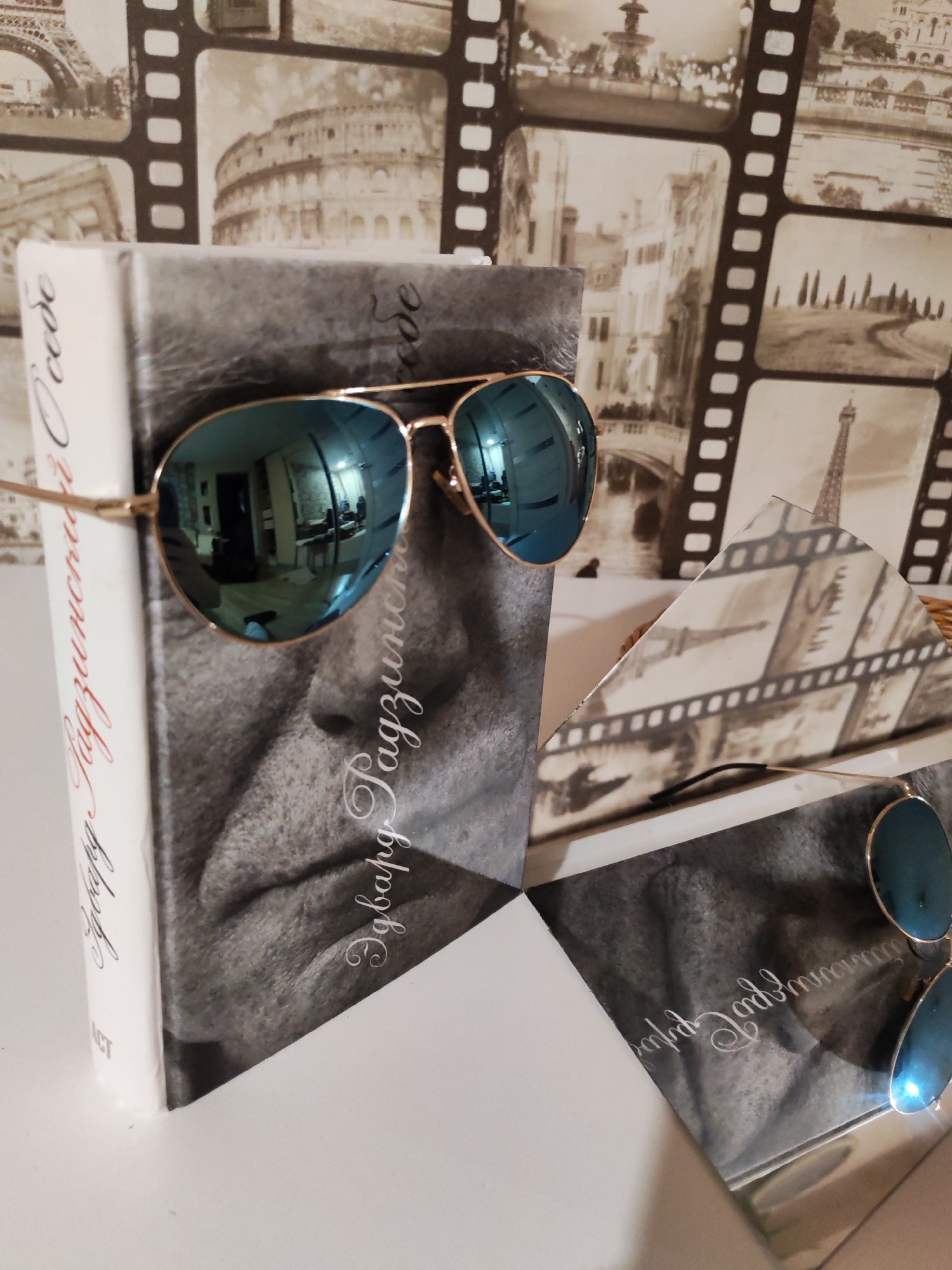 The image shows a book with a black and white portrait of a man on its cover, wearing aviator sunglasses with blue lenses. The book is titled "Eduard Topol". The background features a wallpaper pattern of old movie film reels with famous landmarks. The location, Chaikovsky, Russia, is not directly apparent from the image. The book appears to be placed on a white surface, possibly a table or shelf. In the foreground, there are some other reflective surfaces with the same black and white portrait.